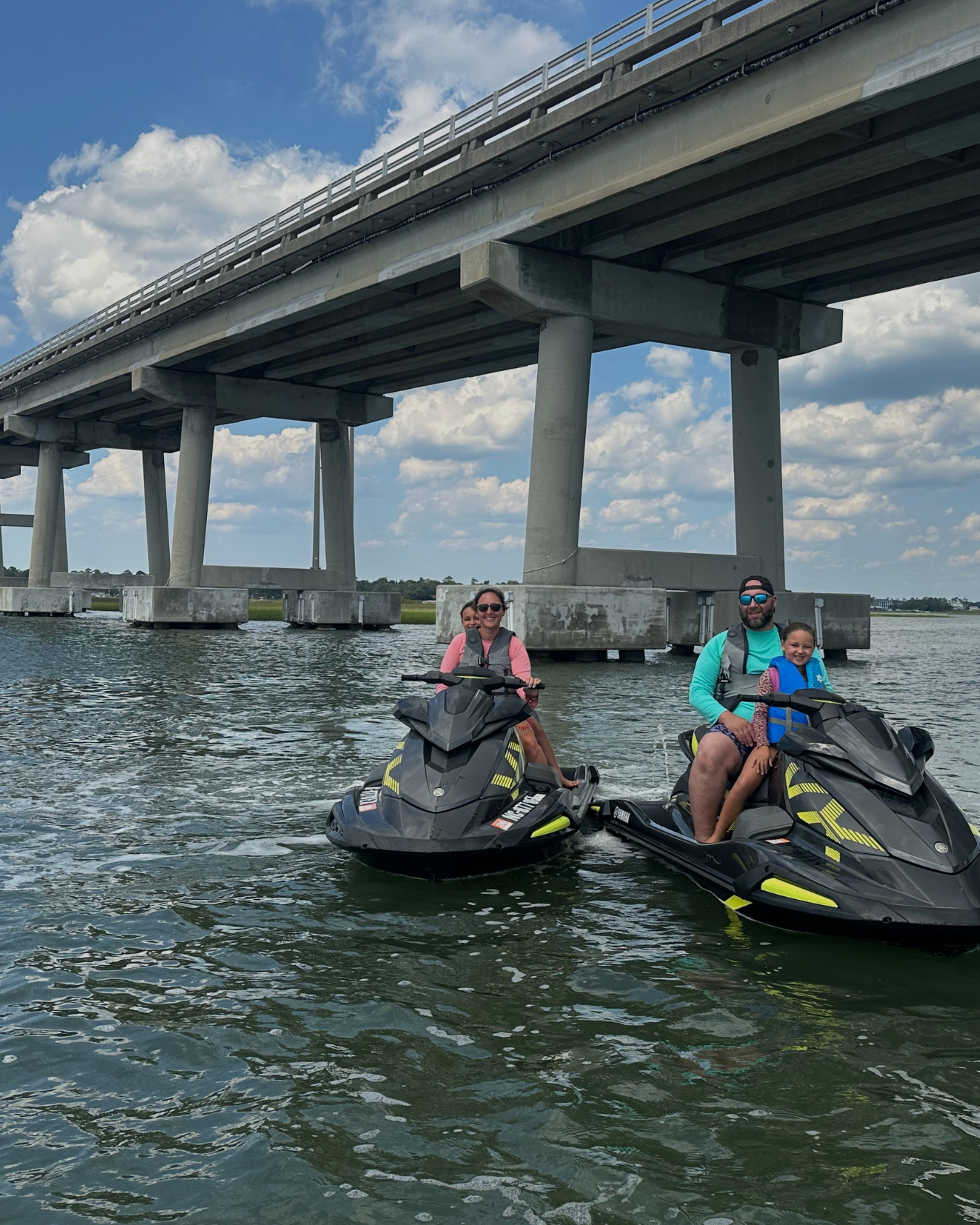 Four children riding three Yamaha jet skis on a sunny day in a waterfront area with houses in the background.