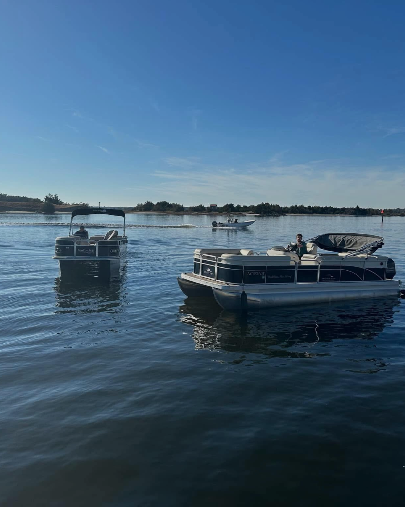 A group of people on a pontoon boat by the water, with blue sky and some clouds, and a shoreline in the background.
