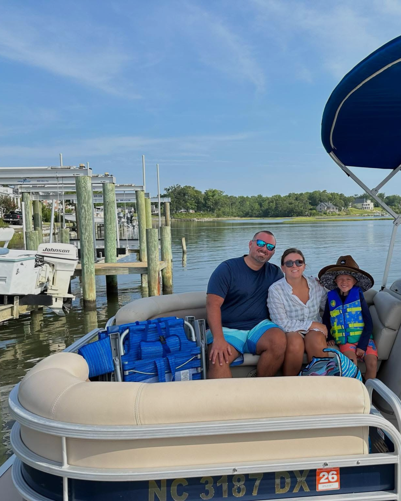 Family and friends on a boat, smiling, enjoying a sunny day on a lake with blue sky and clouds.