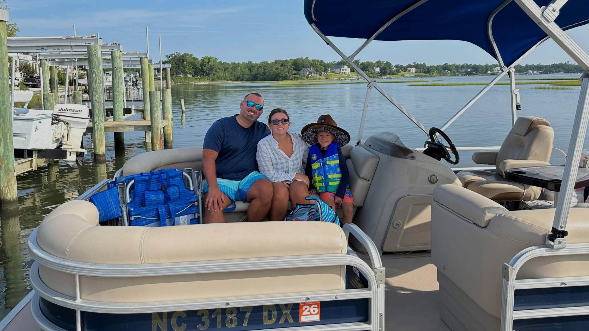 Family and friends on a boat, smiling, enjoying a sunny day on a lake with blue sky and clouds.