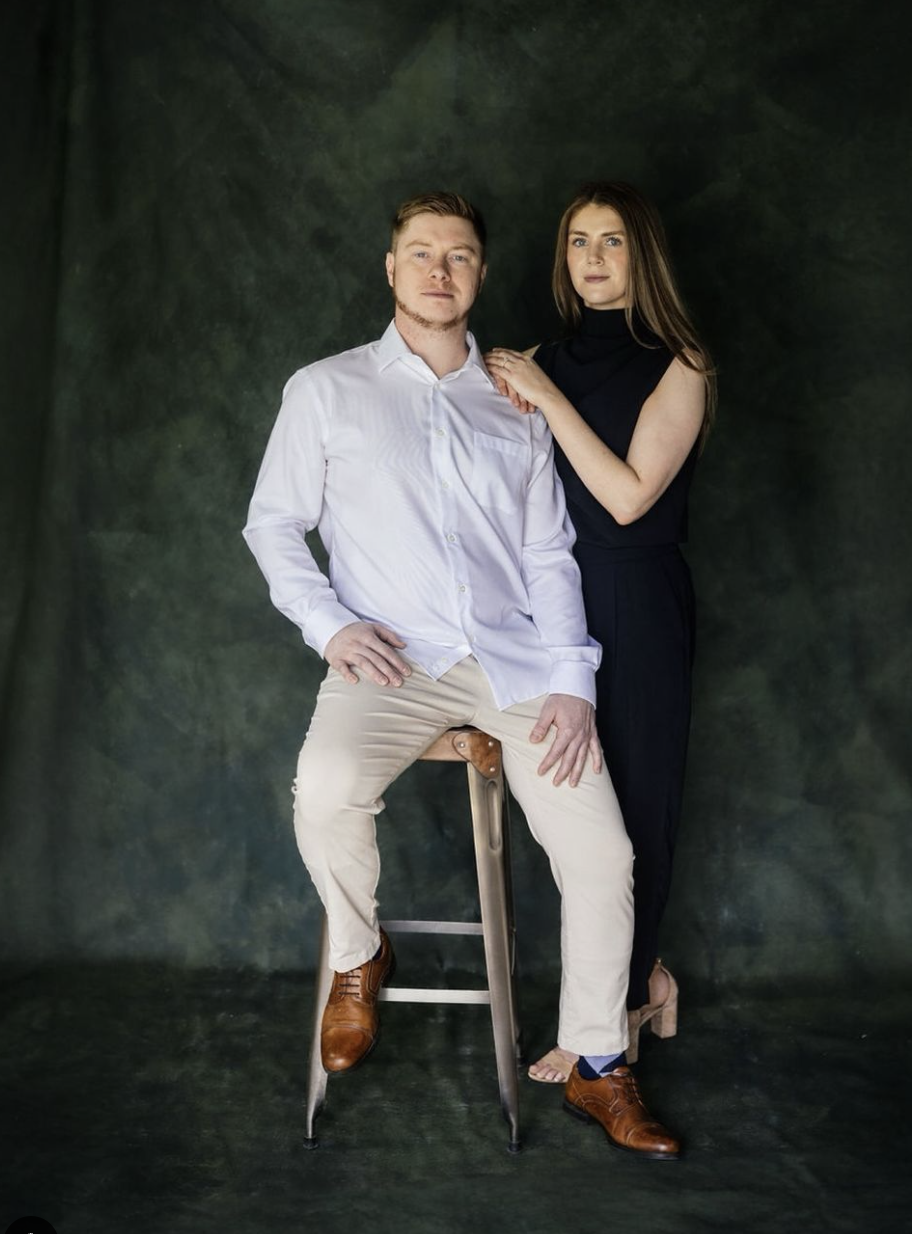A man in a white shirt and beige pants sitting on a stool, with a woman in a black sleeveless top and black pants standing beside him, both posing for a portrait against a dark green background.