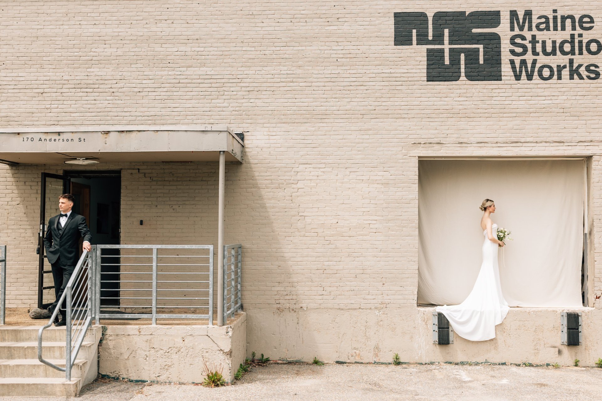 A man in a tuxedo stands on stairs outside a beige brick building, with a woman in a white wedding dress holding a bouquet standing in a window opening covered with a white curtain.
