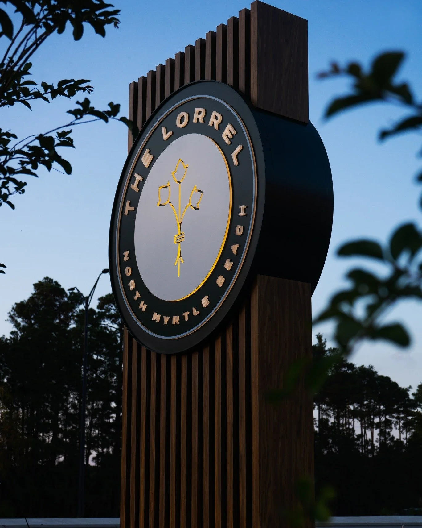 Sign for the Heather Lorrell Township, featuring a stylized flower with three tulips at the center, mounted on a wooden post with a round face, illuminated letters and lights, against a dusk sky with trees in the background.
