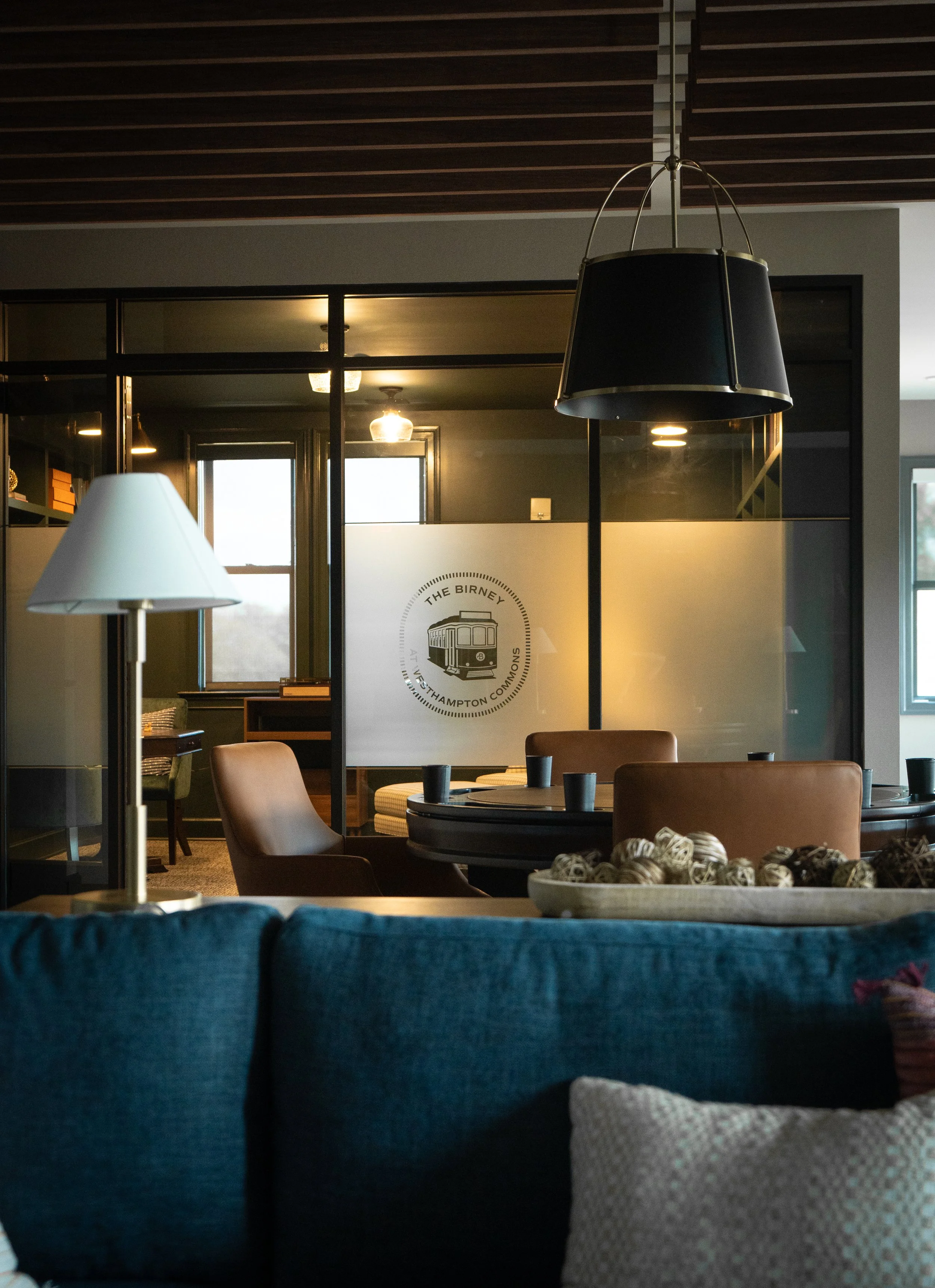 Interior of a modern lounge or common area with pendant lights, a lamp, wooden chairs, tables, and a decorative bowl filled with yarn balls, with a frosted glass wall featuring a logo of a tram and the text 'The Birney at Southampton Common'.