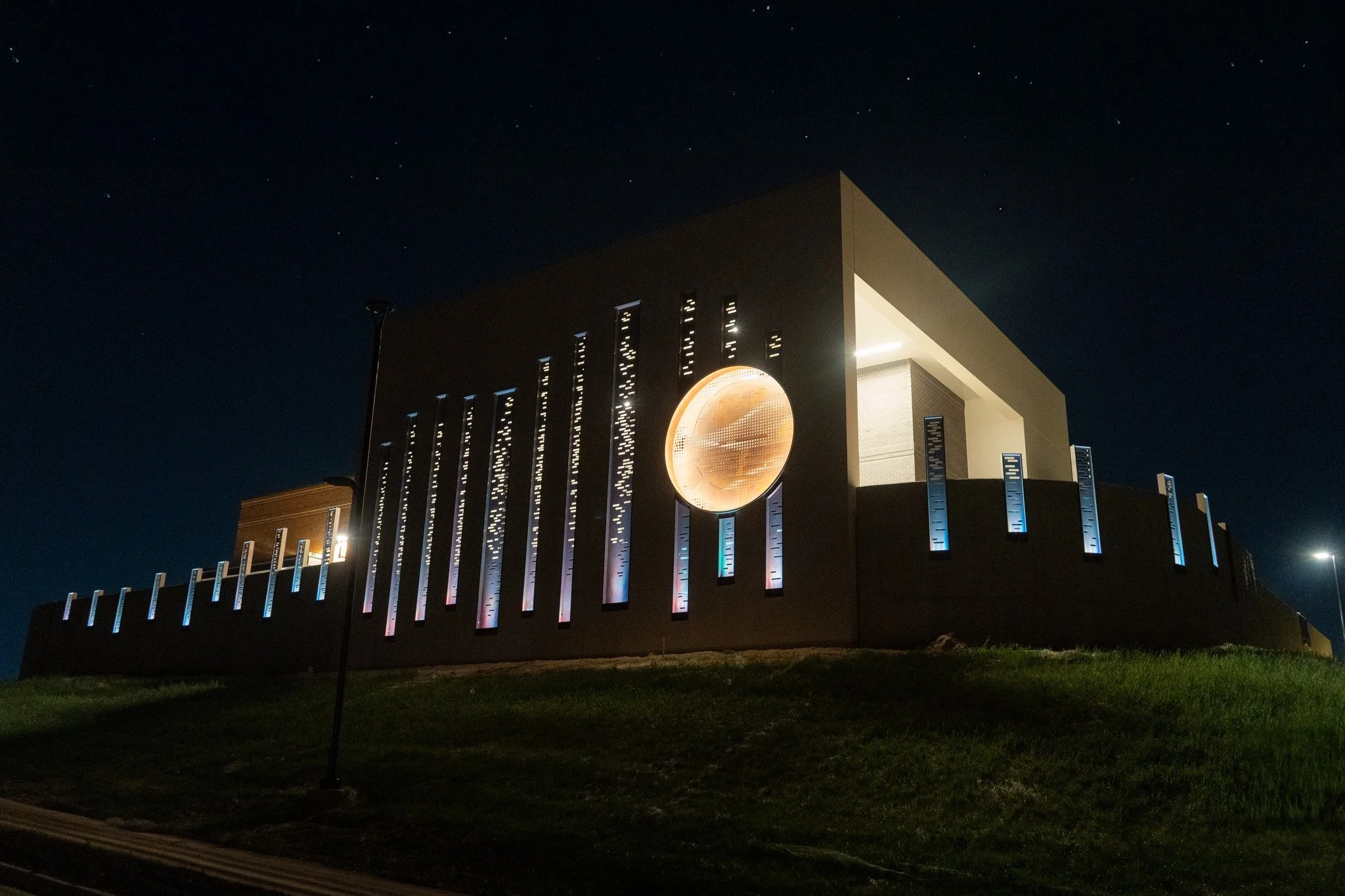 Night view of a modern building with vertical light strips, a large circular illuminated sculpture, and a well-lit open area, set against a dark sky with stars.