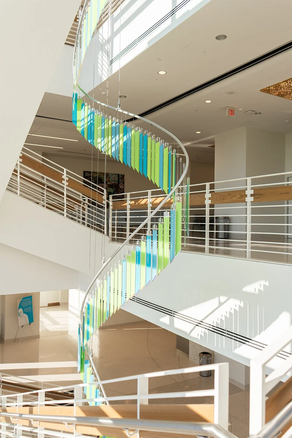 Interior view of a modern building with a colorful, spiraling staircase decorated with hanging panels in shades of blue, green, and yellow.
