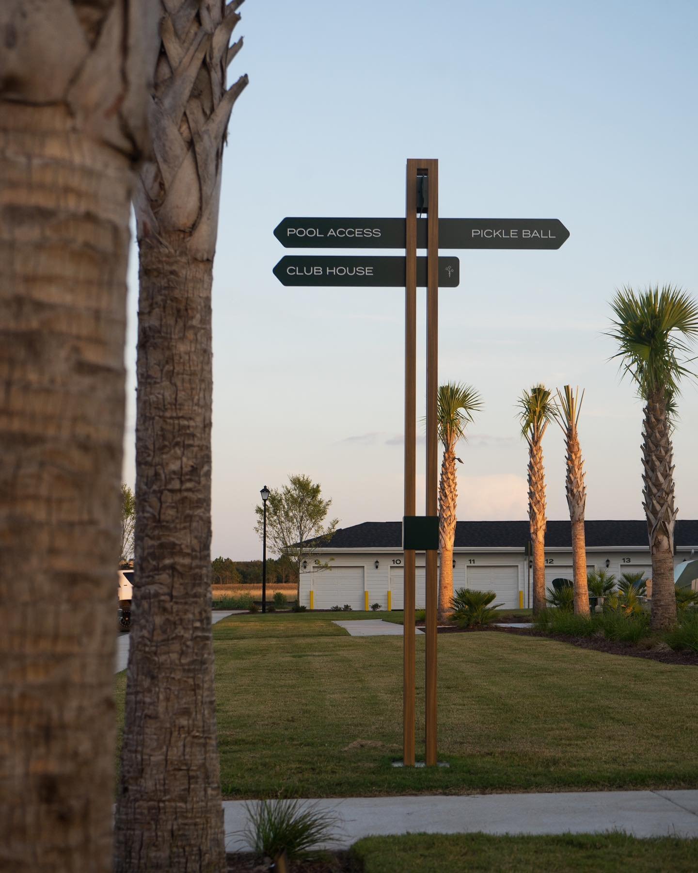 A signpost with directions to pool access, pickleball, and club house in a tropical residential area with palm trees and a garage in the background at sunset.