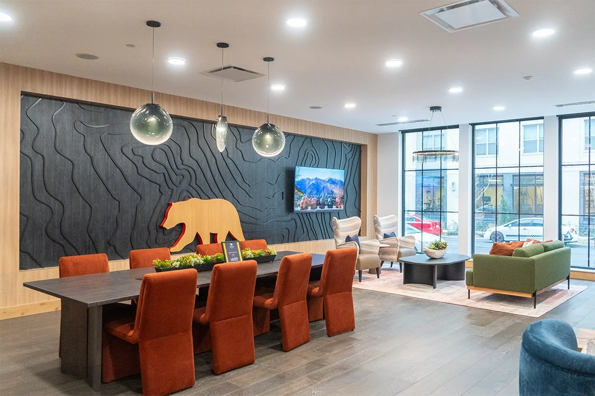Modern lounge area with a large black wall featuring a carved bear silhouette, a flat-screen TV, a long table with eight orange chairs, a seating area with a green couch, two cream armchairs, and a black coffee table. Large windows overlook cars park