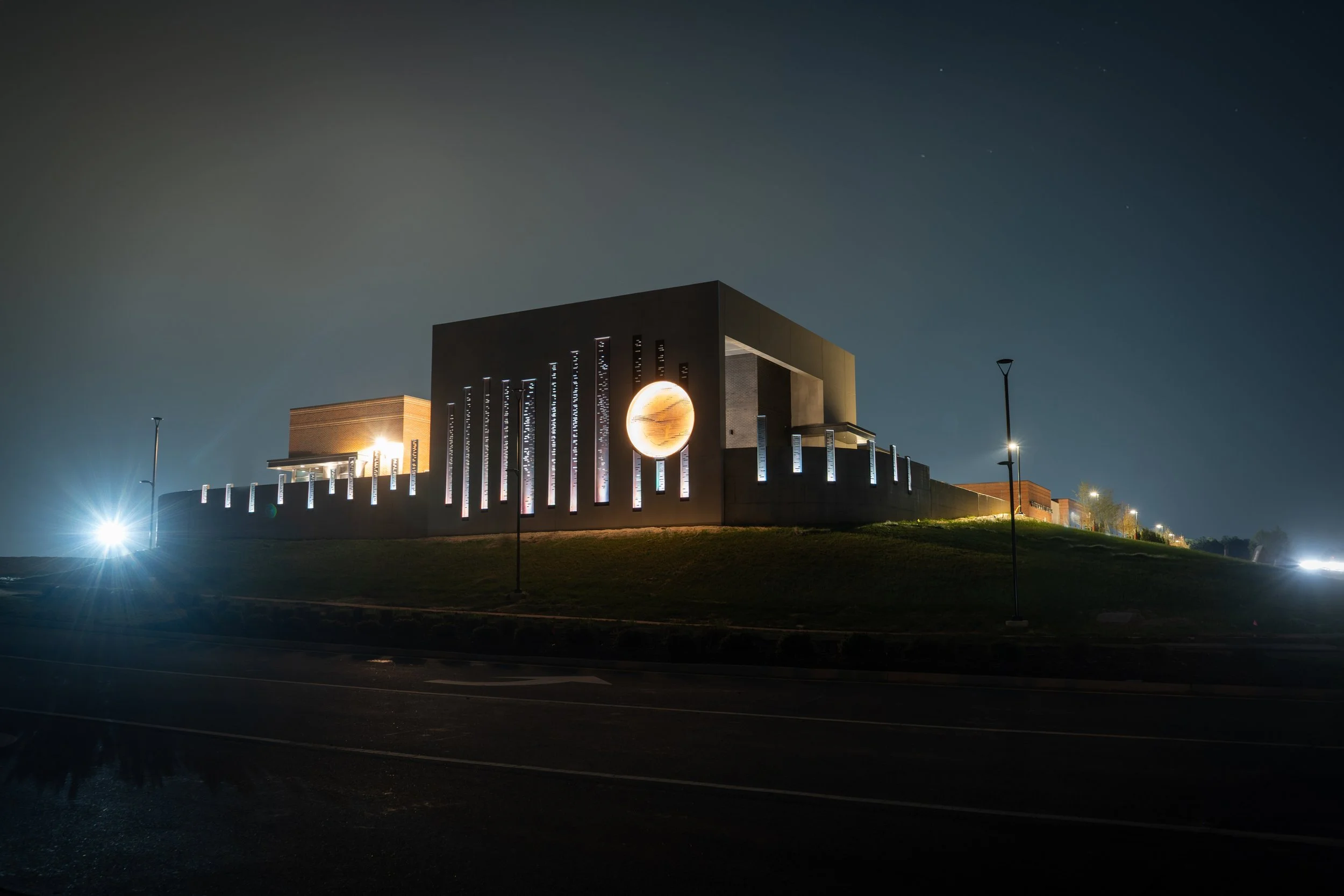 Night view of a modern building with a large illuminated globe and vertical light fixtures, on a grassy hill next to a road with streetlights.