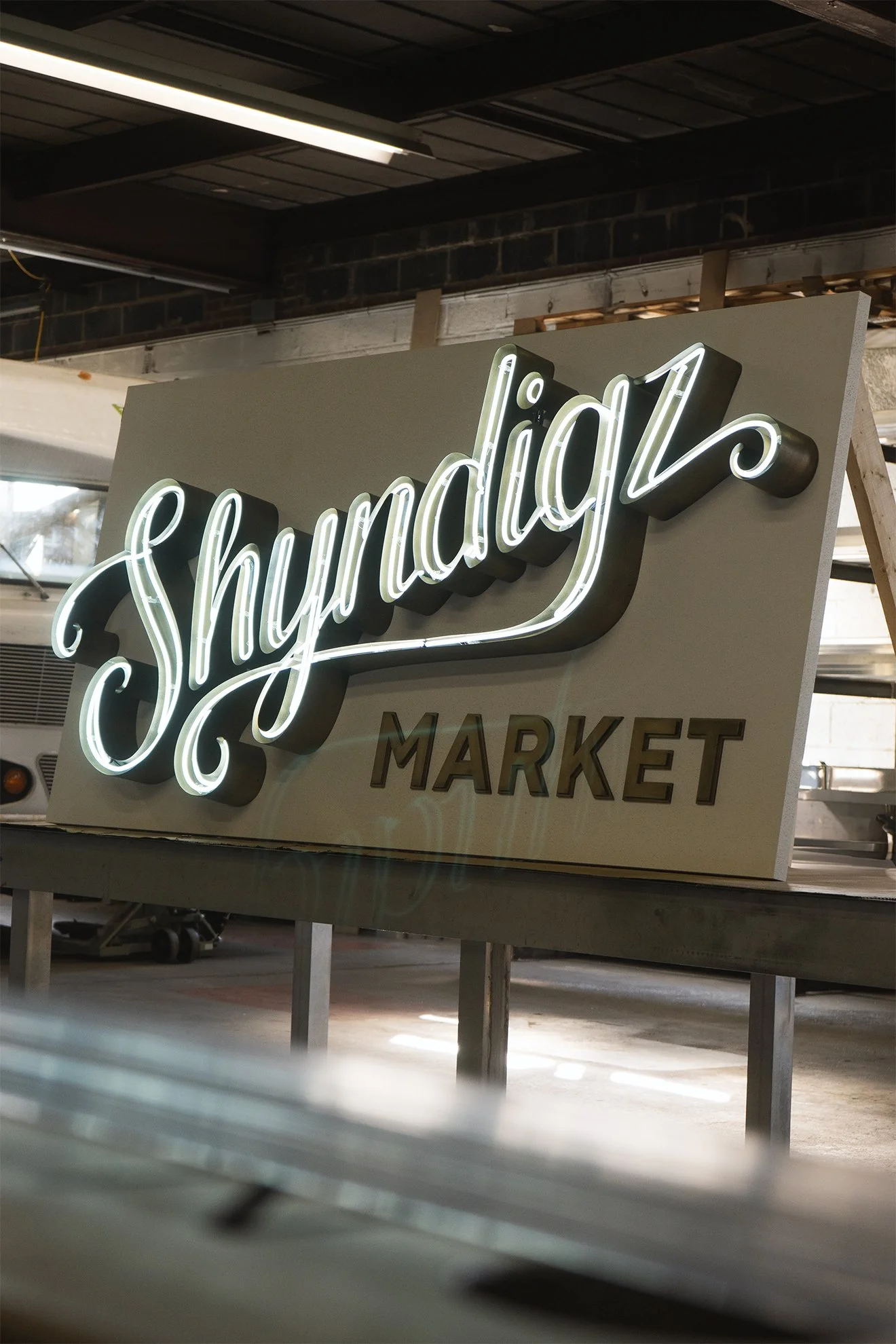 Neon sign reading 'Shyndigz MARKET' inside a store with industrial interior, including a brick wall and metal ceiling beams.