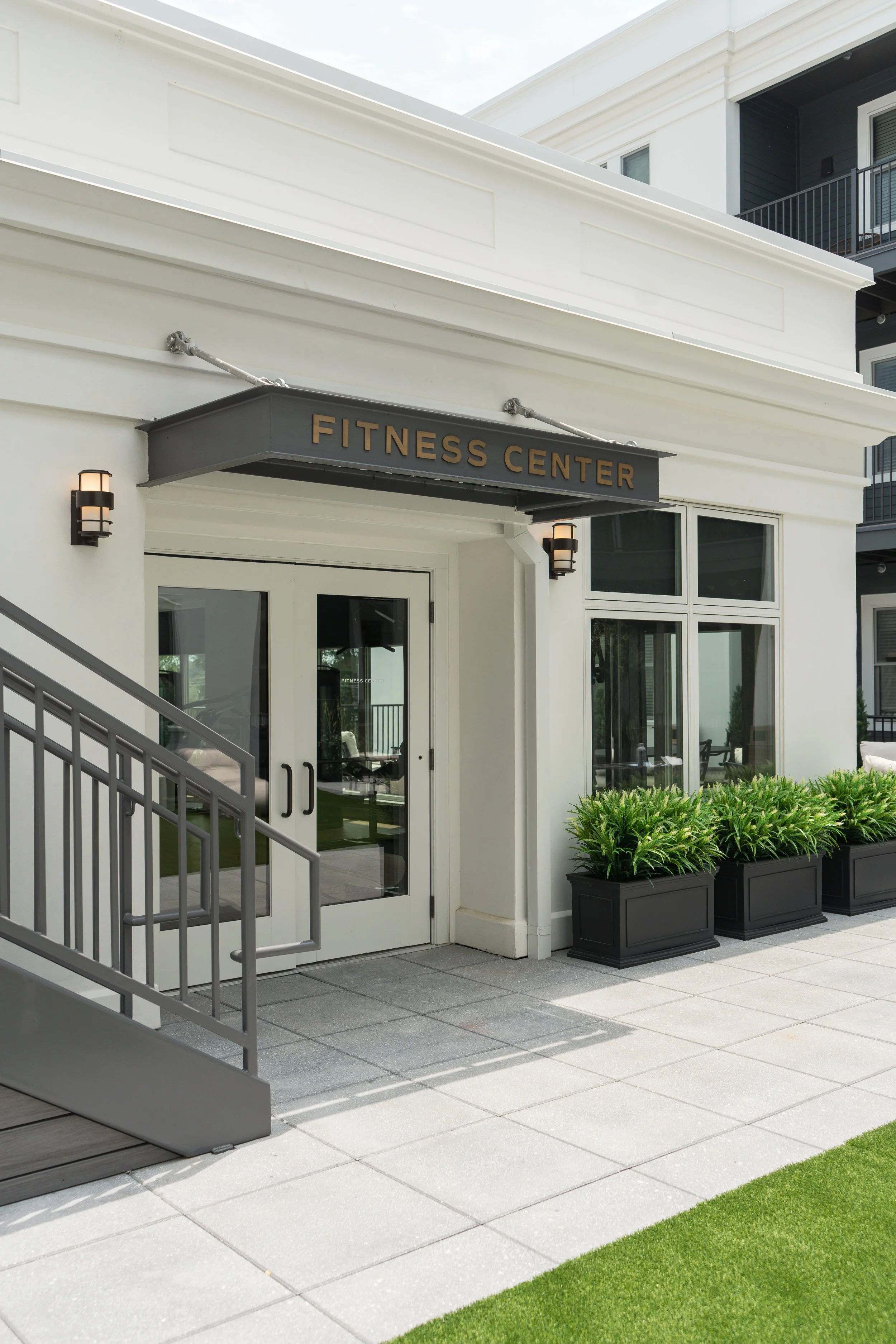Exterior view of a fitness center with a white building, black sign reading 'Fitness Center', glass door, and large windows. Black planters with green plants outside, and a metal staircase to the left.