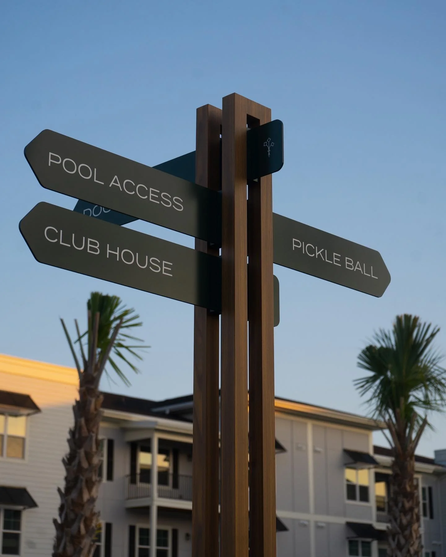Signpost with four directional signs pointing to pool access, club house, pickle ball, and an illegible sign, in front of residential buildings and palm trees under a clear sky.