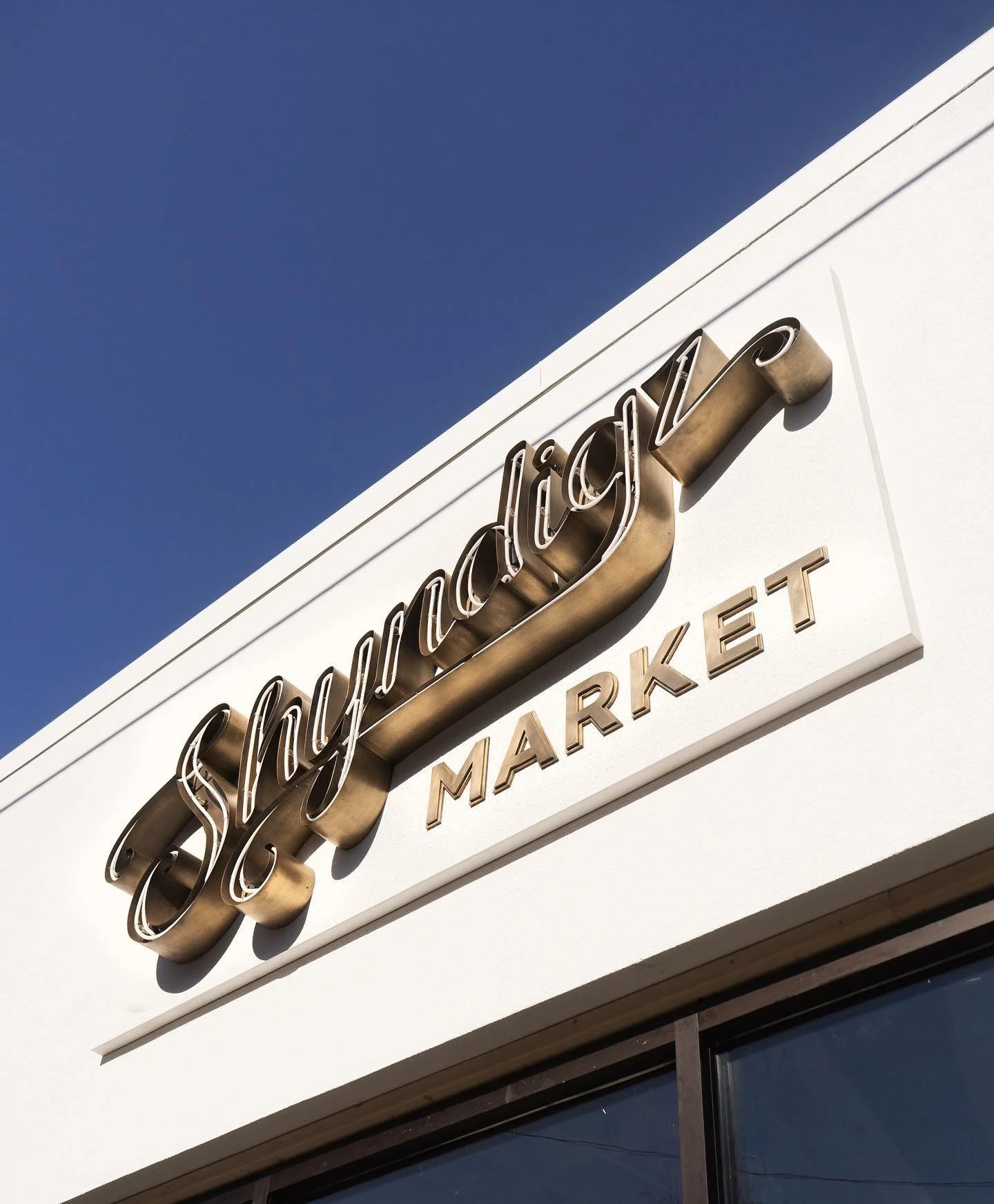 Sign for a Shmayz Market with gold lettering on a white background under daylight.