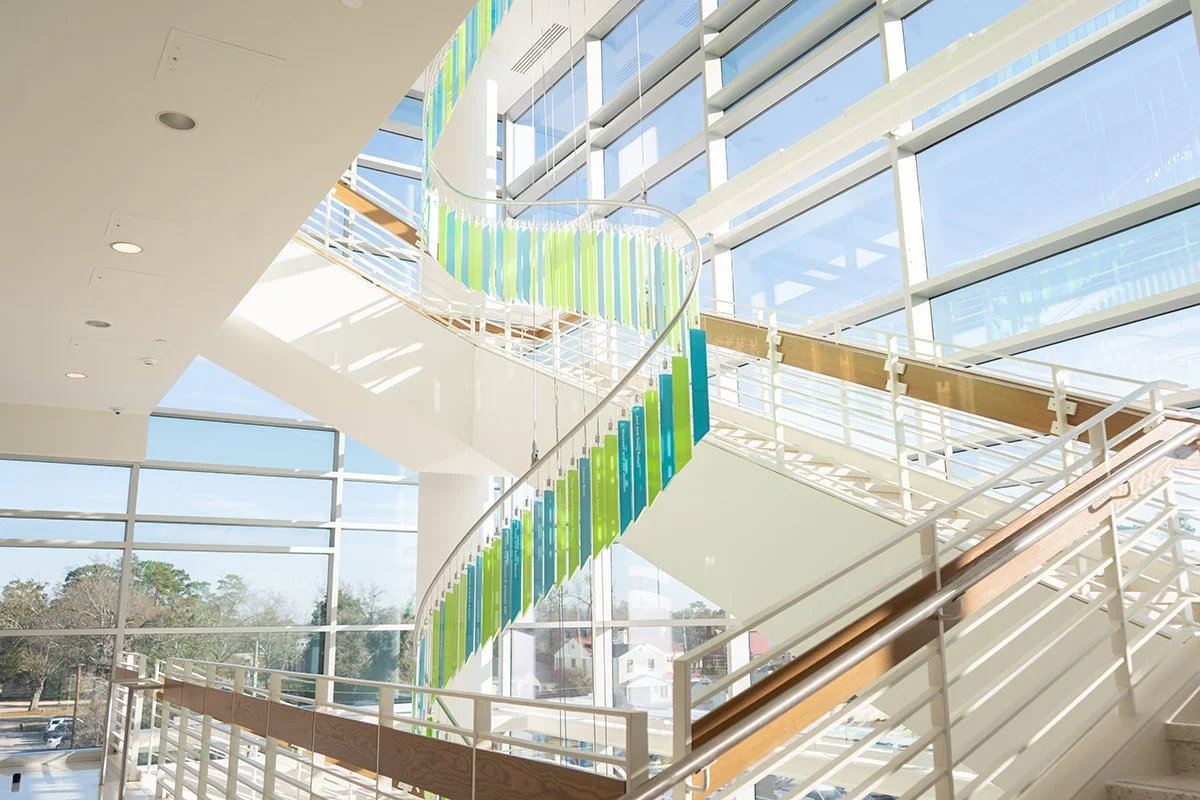 Modern indoor staircase with glass walls and colorful vertical art installation, leading to an upper floor in a well-lit building.
