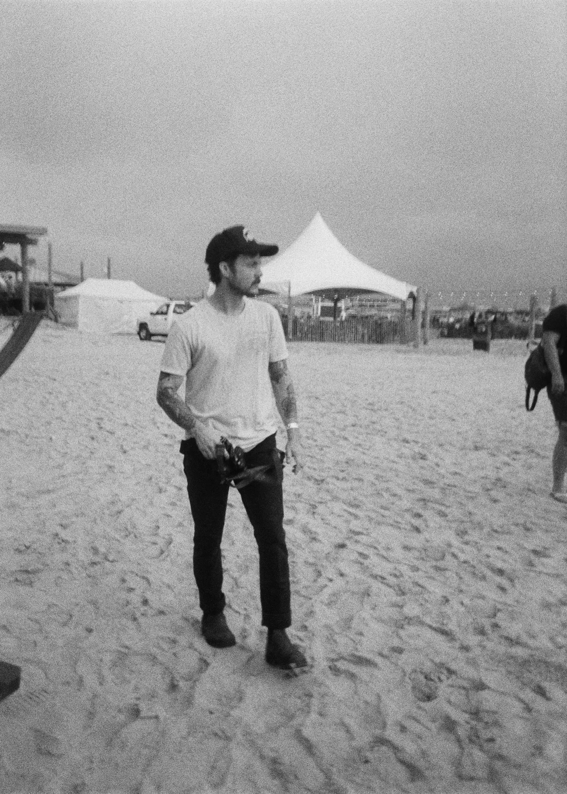 A man standing on a sandy beach holding a camera, wearing a cap, a t-shirt, and jeans, with tents and beach structures in the background.