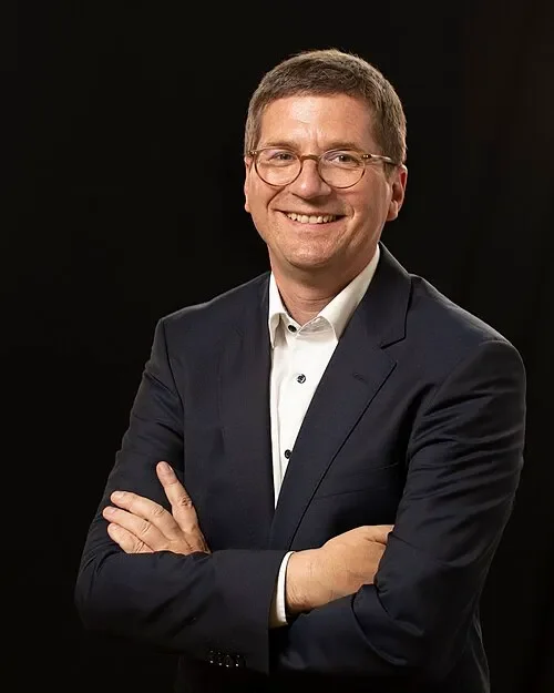 A smiling man in a dark suit, white shirt, and glasses, posing with crossed arms against a black background.