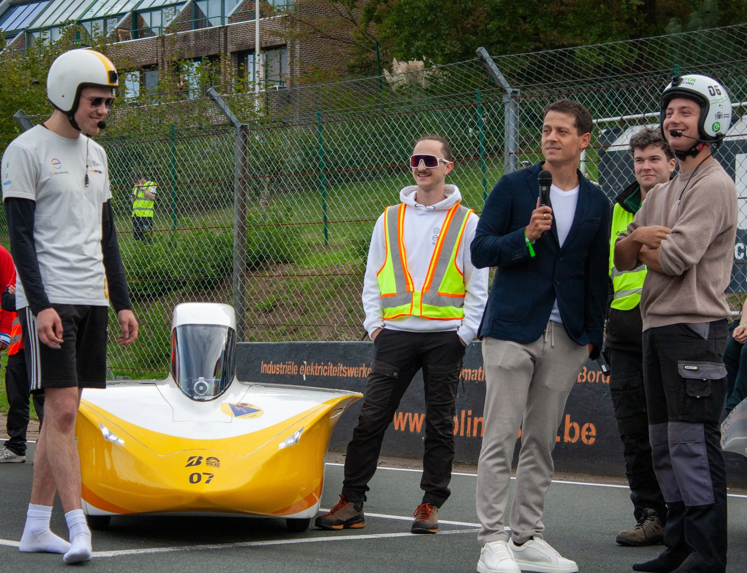 Group of people at a racing event, some wearing helmets and safety vests, around a yellow race car.