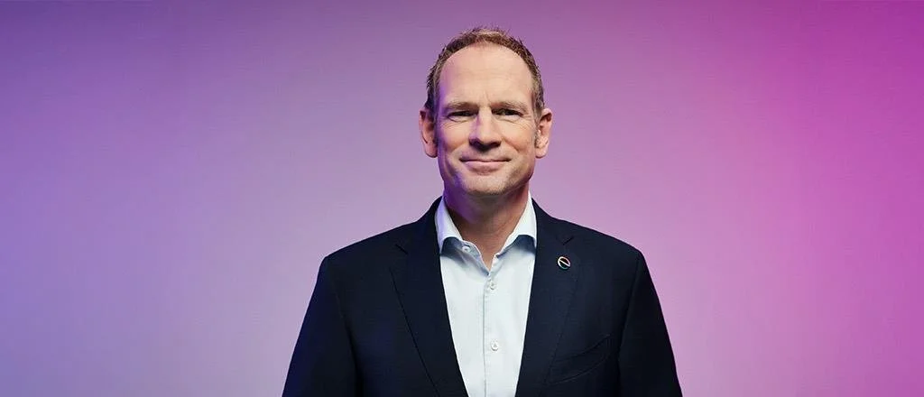 Portrait of a middle-aged man in a dark suit and white shirt, smiling slightly, against a purple gradient background.