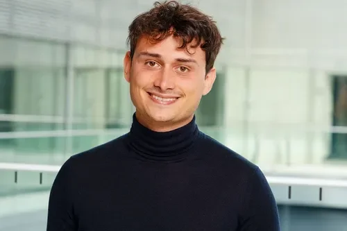 Portrait of a young man with curly brown hair, wearing a black turtleneck sweater, smiling in front of a modern glass building.