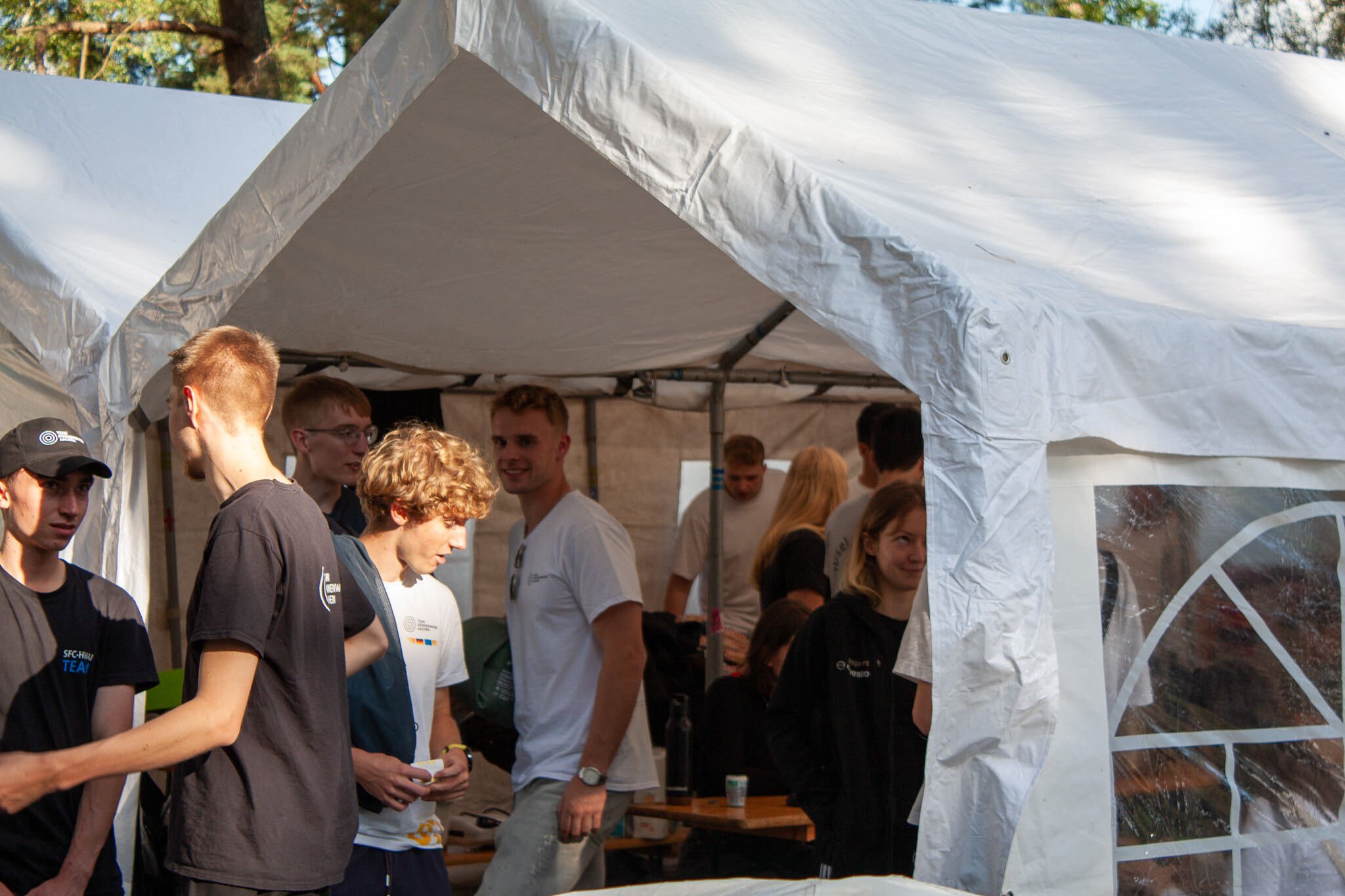 A group of young people gathered under a white tent outdoors in a park or garden setting.