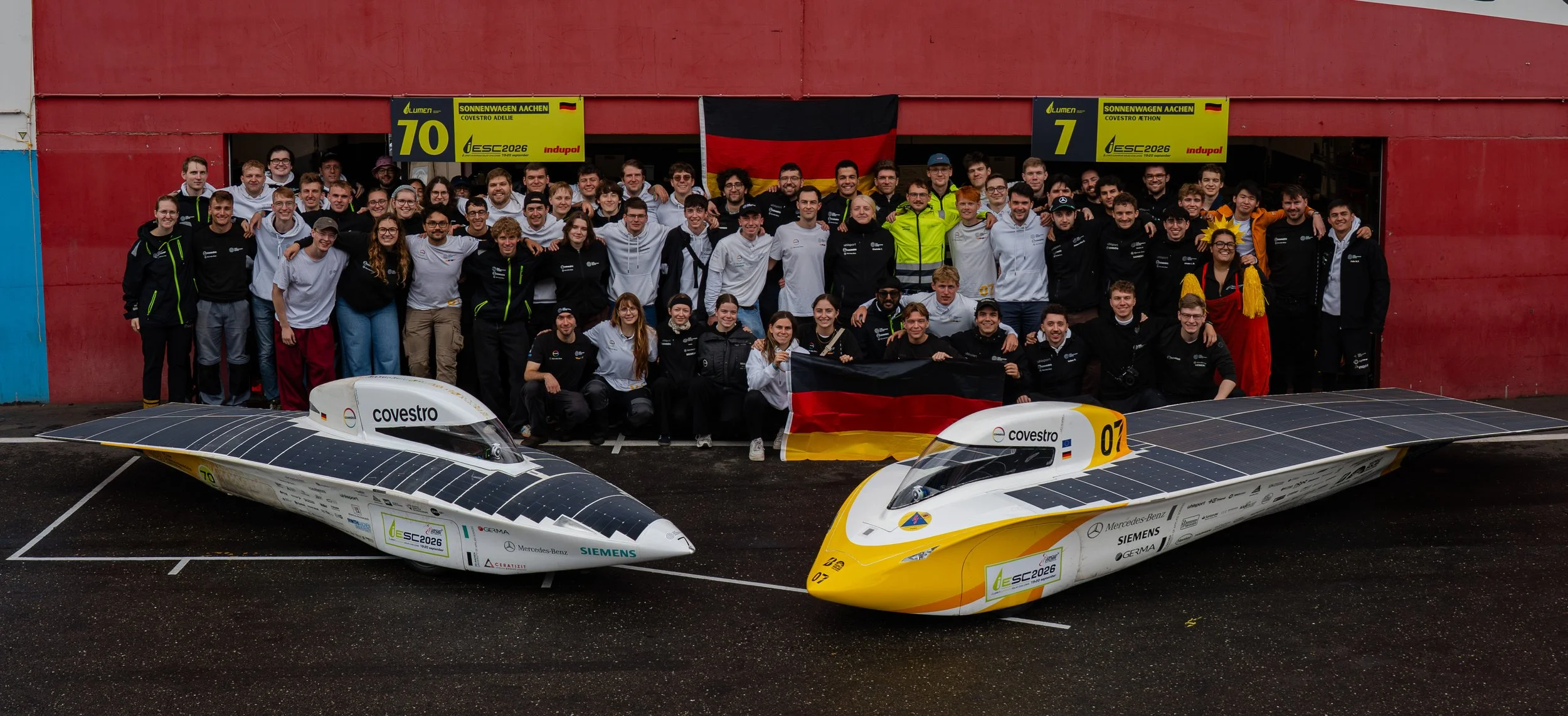 A group of people gathered in front of a red building, posing with two solar-powered racing vehicles, with German flags displayed behind them. The vehicles are sleek, solar-powered race cars, marked with numbers 70 and 07, and sponsor logos.
