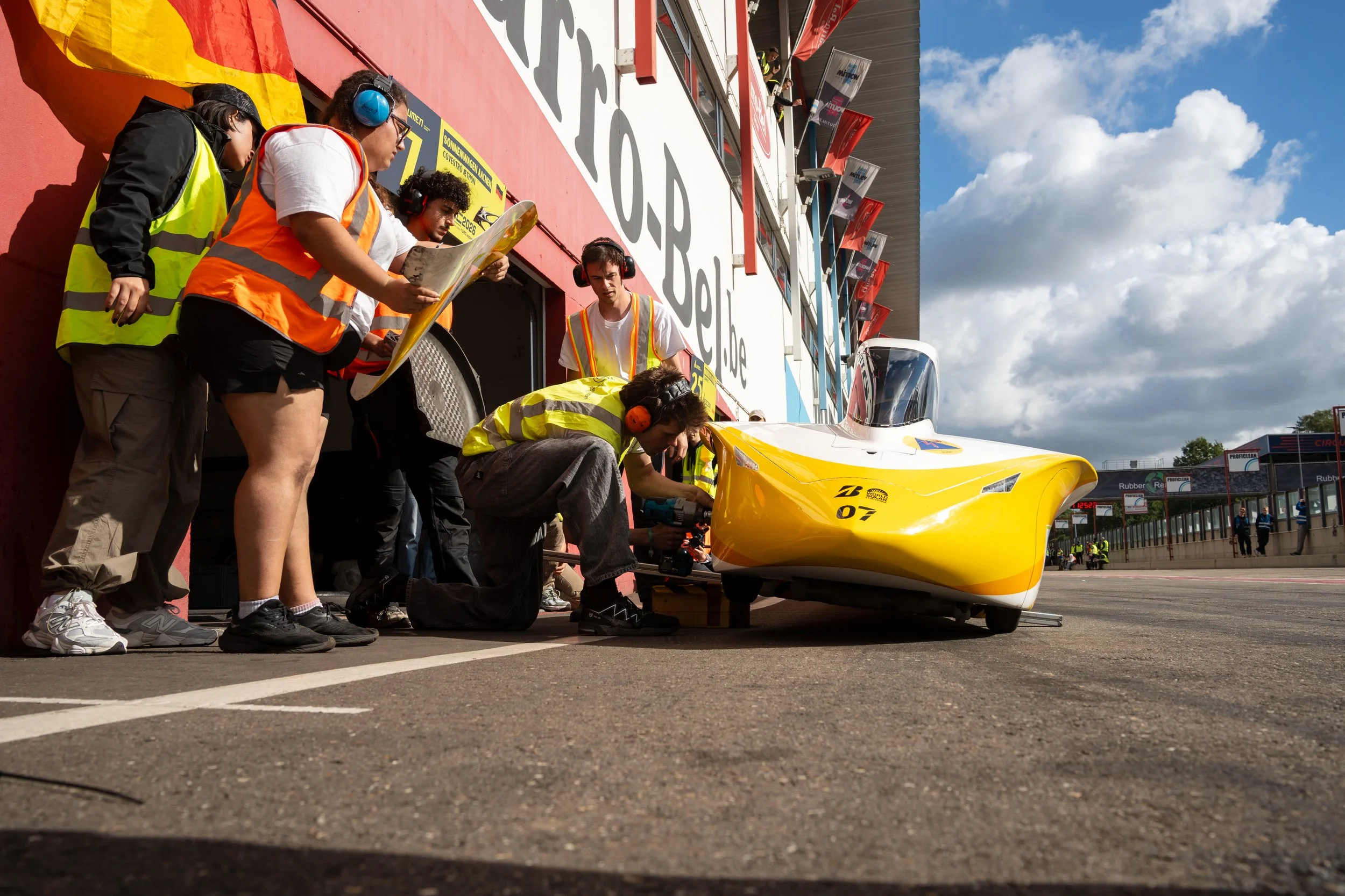 Race team working on a yellow racing car in the pit lane, with team members wearing safety vests and ear protection, and a grandstand with spectators in the background.