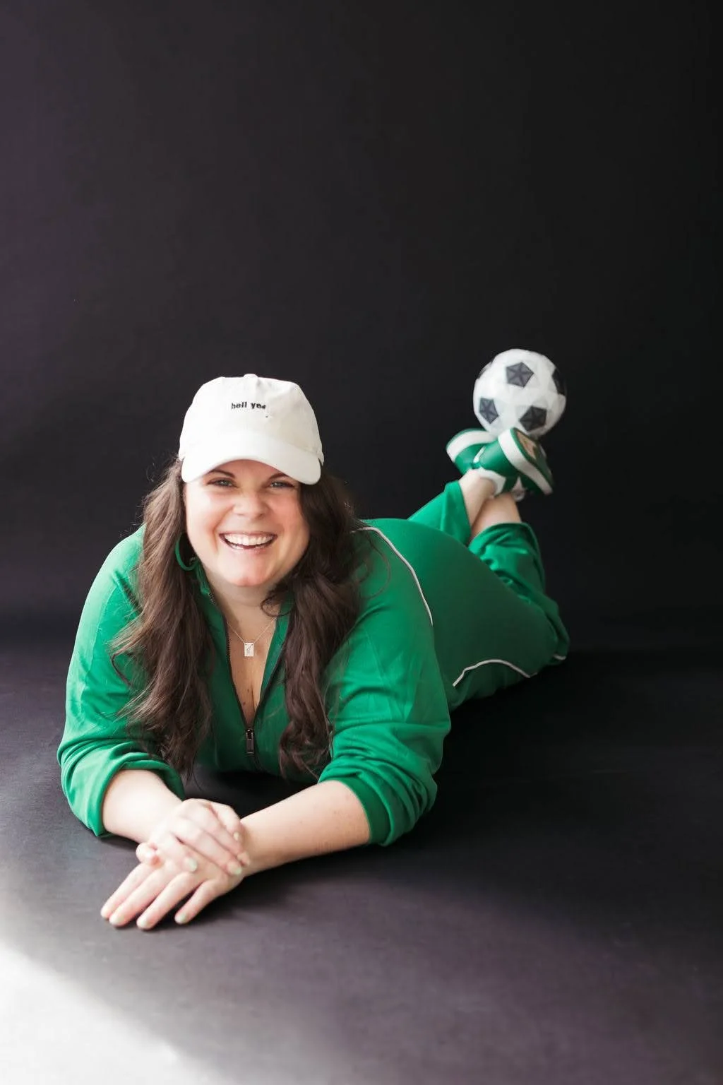 Woman lying on her stomach on a black surface, smiling, wearing a white cap and a green sports jacket, with one foot raised wearing a sports shoe and holding a soccer ball.