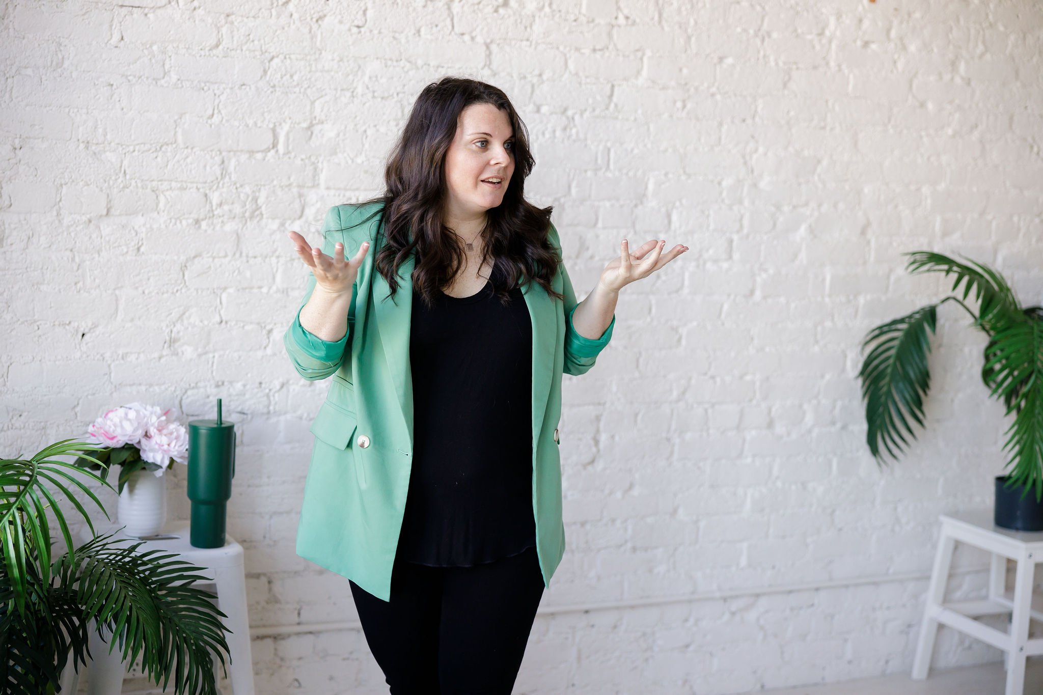 Woman with dark hair, wearing a green blazer and black top, gesturing with her hands in a room with white brick wall, plants, and flowers.