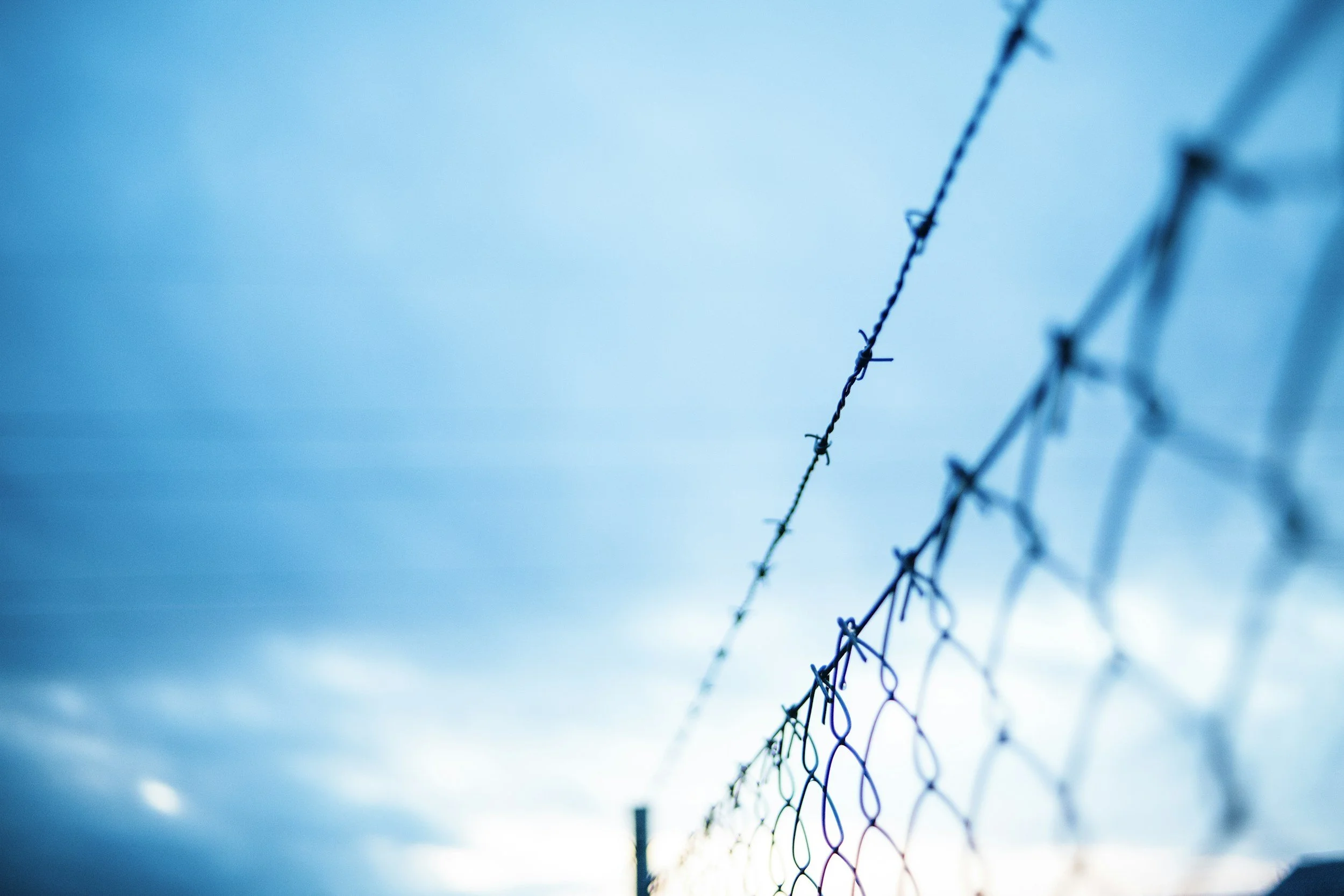 Close-up of a chain-link fence with barbed wire on top, against a cloudy blue sky.