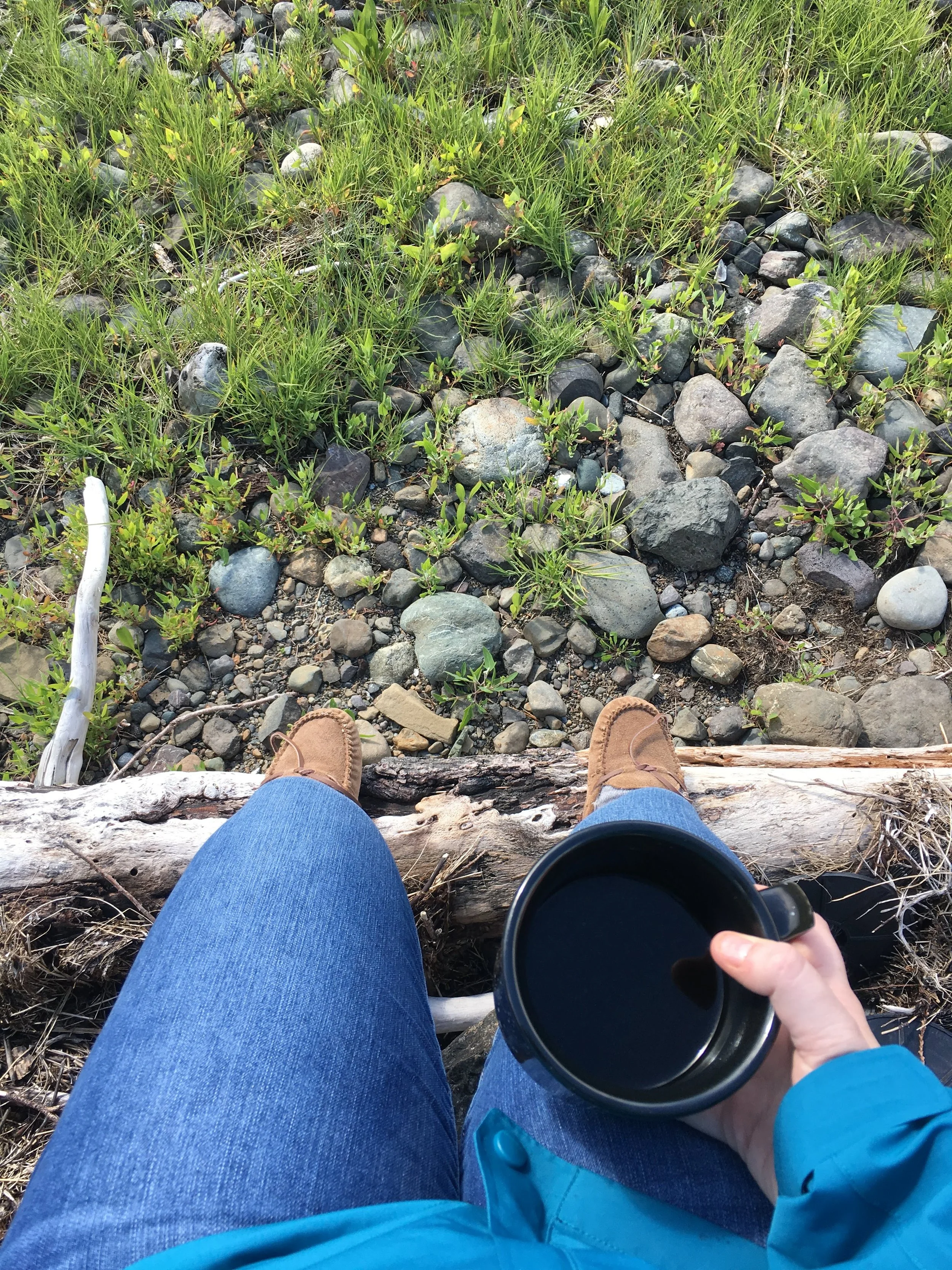 Person sitting outdoors on a log, wearing blue jeans and brown shoes, holding a black mug filled with a dark beverage, overlooking a rocky and grassy area.
