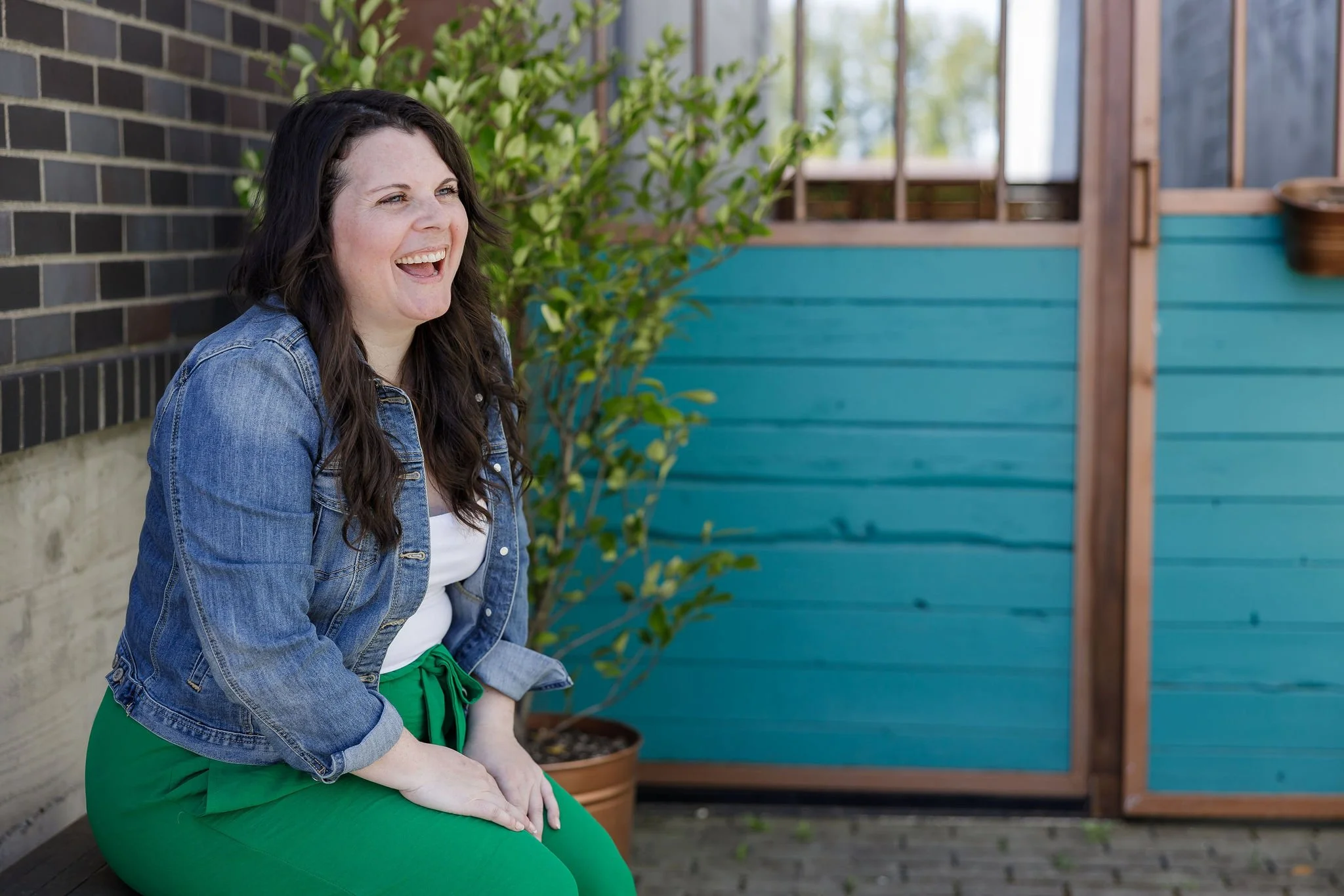 A woman with long dark hair wearing a denim jacket and green pants is sitting outside on a wooden deck, laughing.