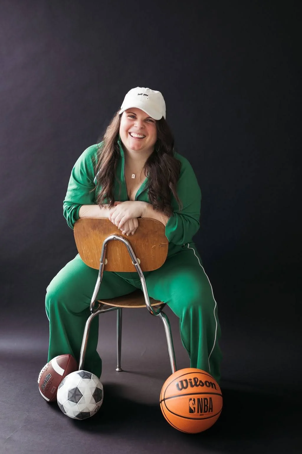 A young woman with long dark hair wearing a white cap, a green tracksuit, and red sneakers, smiling and sitting on a wooden chair. She is leaning forward on the chair's backrest with her arms crossed. There are sports balls at her feet, including a f