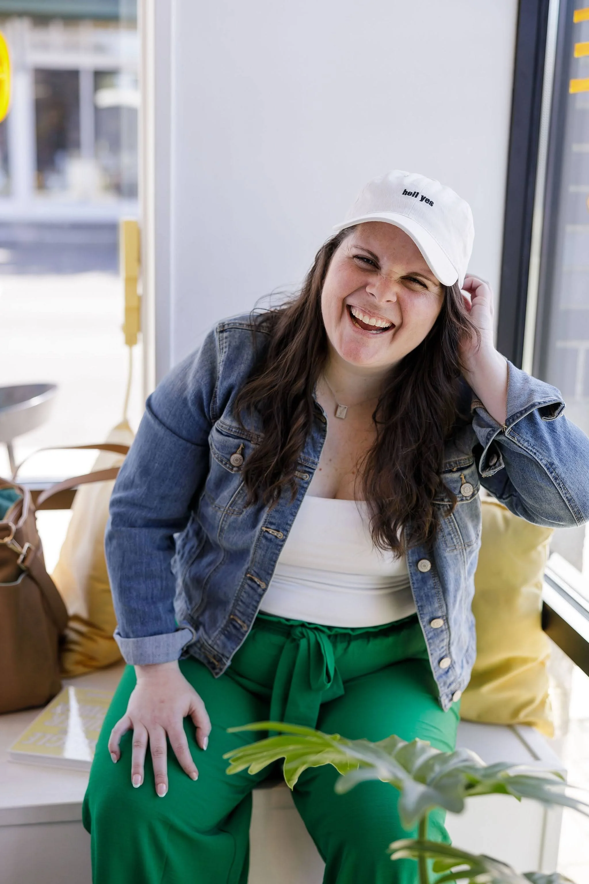 A woman smiling and squinting at the camera, sitting by a window, wearing a white cap, denim jacket, white top, and green pants. Custom Notion business operating system dashboard for high-capacity founders.