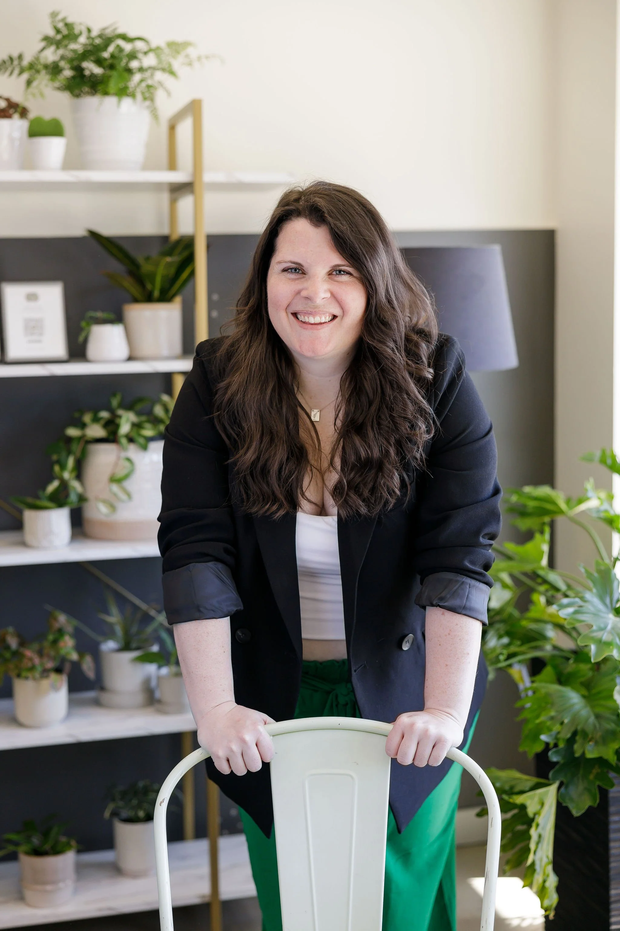 A woman with long, wavy brown hair smiling and leaning on a white chair in a room with green plants and a black and white shelf.
