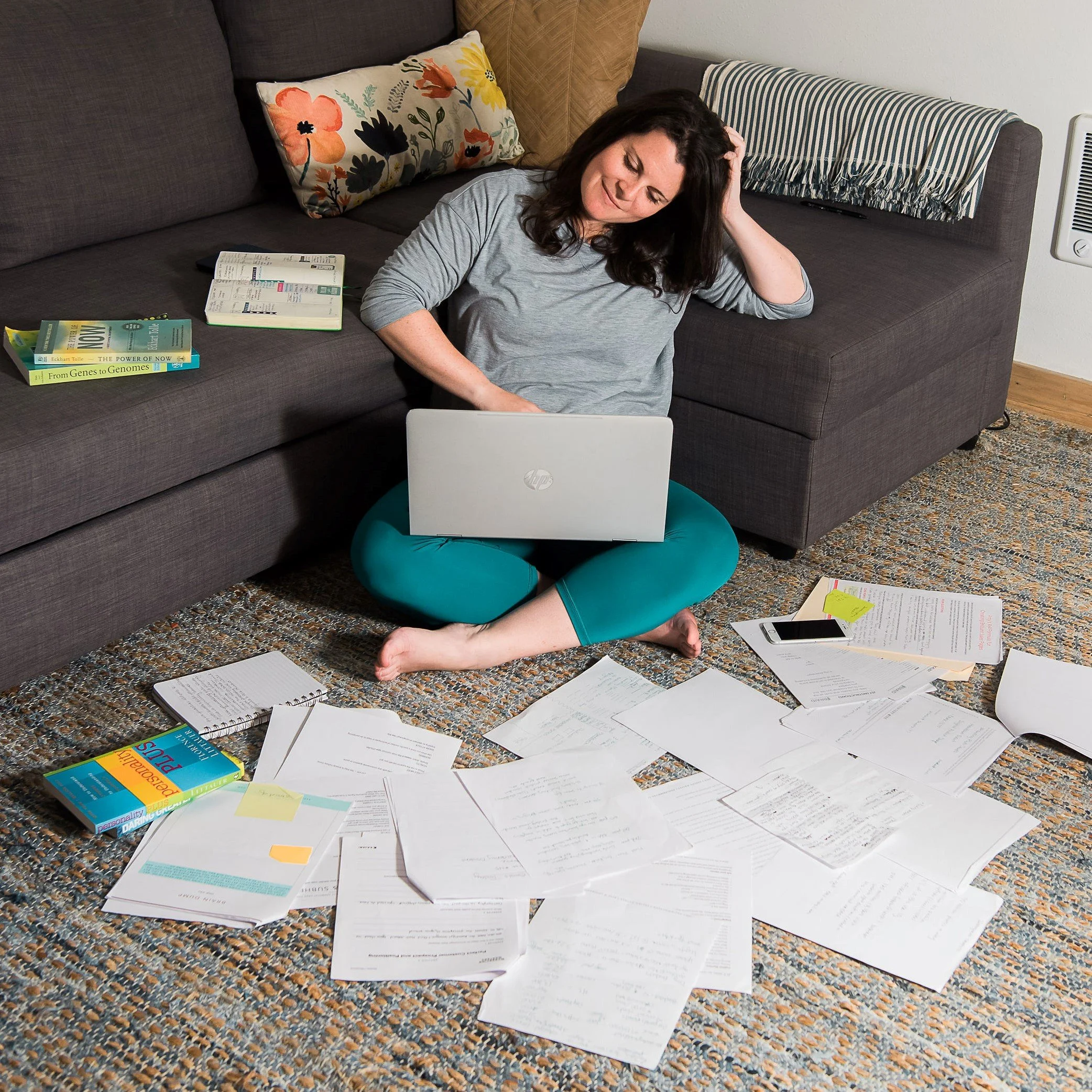 Woman sitting cross-legged on the carpeted floor, working with a laptop surrounded by scattered papers, notebooks, books, a smartphone, and sticky notes, with a sofa and cushions in the background.