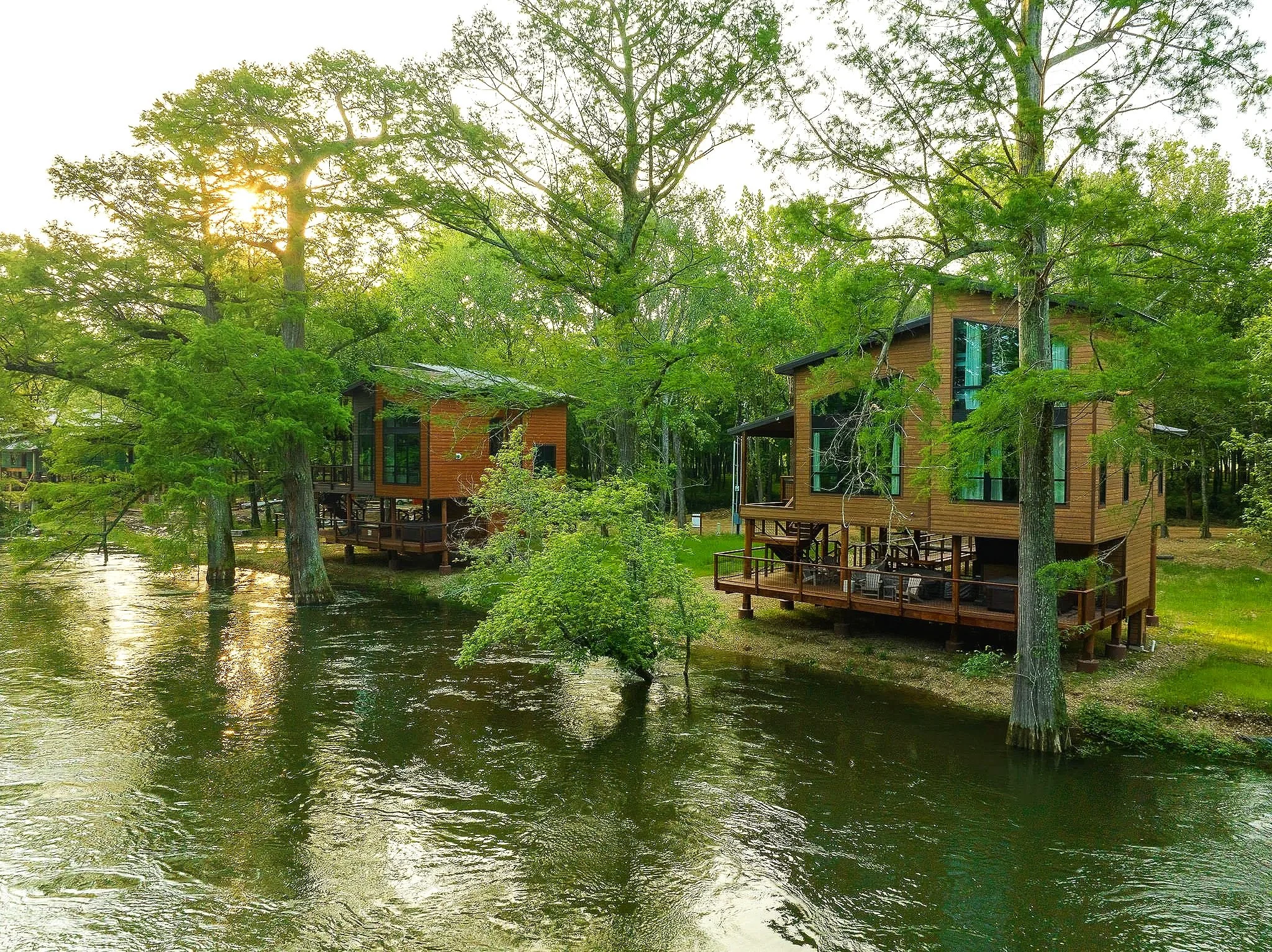 A serene scene with several wooden houses built on stilts beside a calm river, surrounded by tall green trees with sunlight shining through the branches.