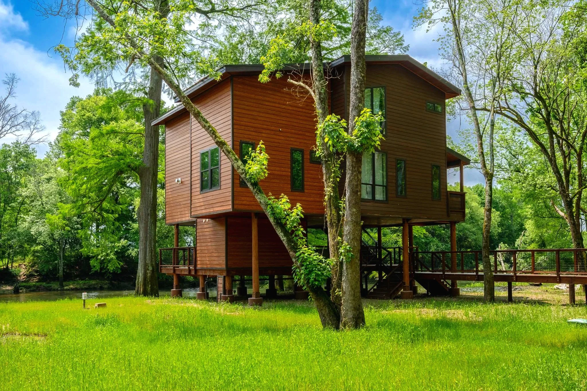 A modern wooden house elevated on stilts amidst green trees with a lake in the foreground and blue sky with clouds in the background.