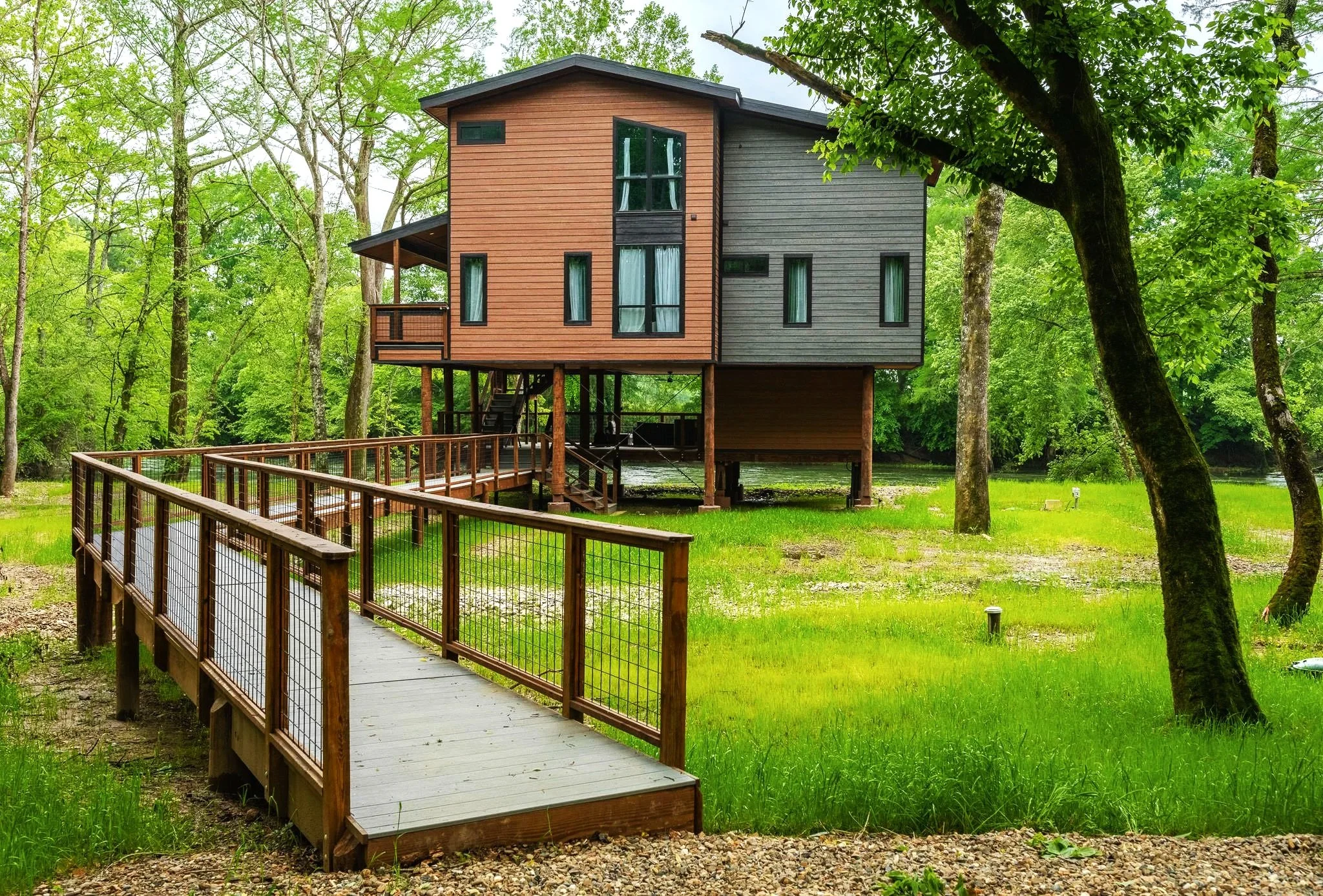 A modern two-story house with alternating brown and gray wooden siding, elevated on stilts, surrounded by green trees and grass, with a wooden walkway leading to the entrance.