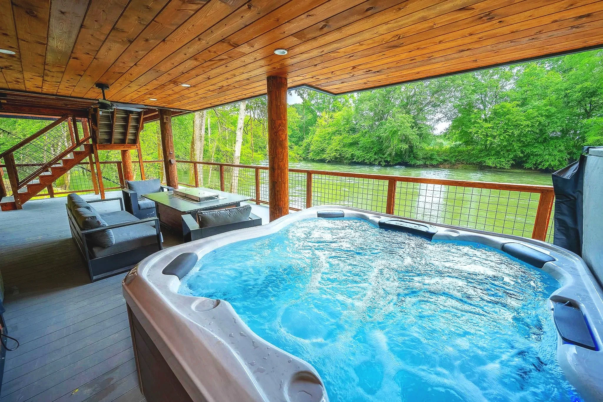 Covered deck with hot tub, patio seating area with chairs and a table, overlooking a lake and green trees.