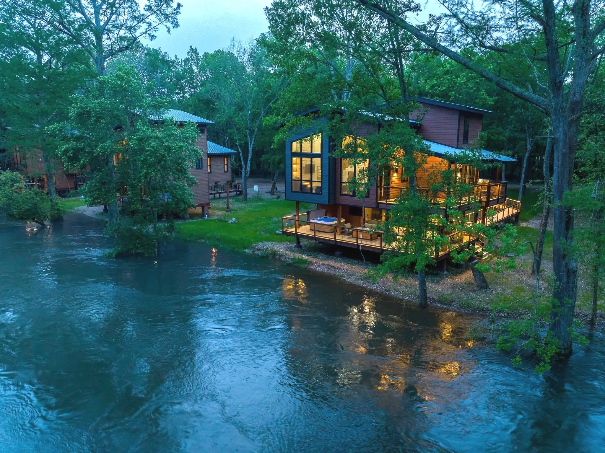 A view of a house built among trees near a body of water, with warm lights glowing from inside the house, and the surrounding trees reflecting in the water during early evening.