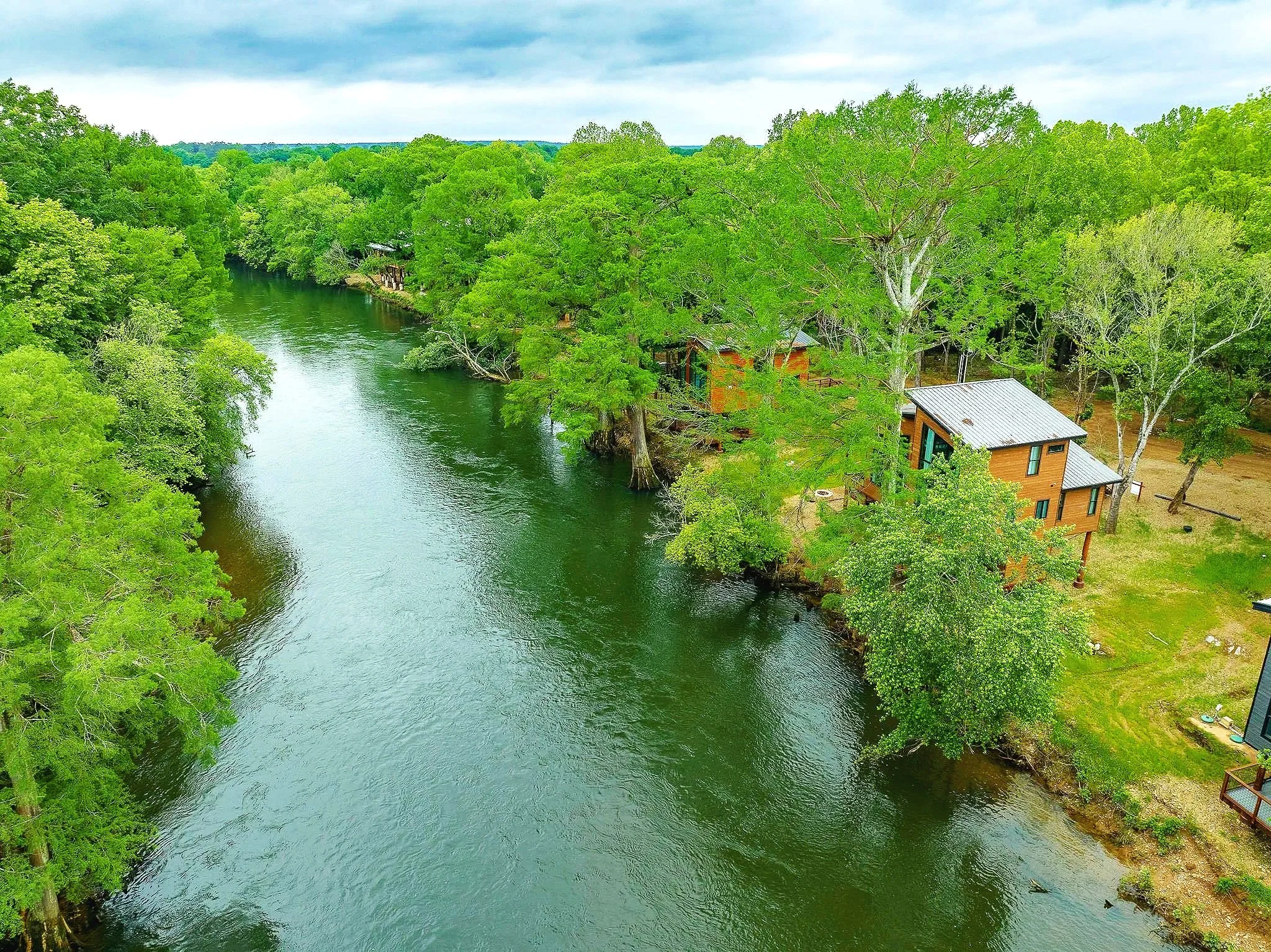 Aerial view of a green river surrounded by dense trees and a few small cabins with metal roofs along the shoreline under a cloudy sky.