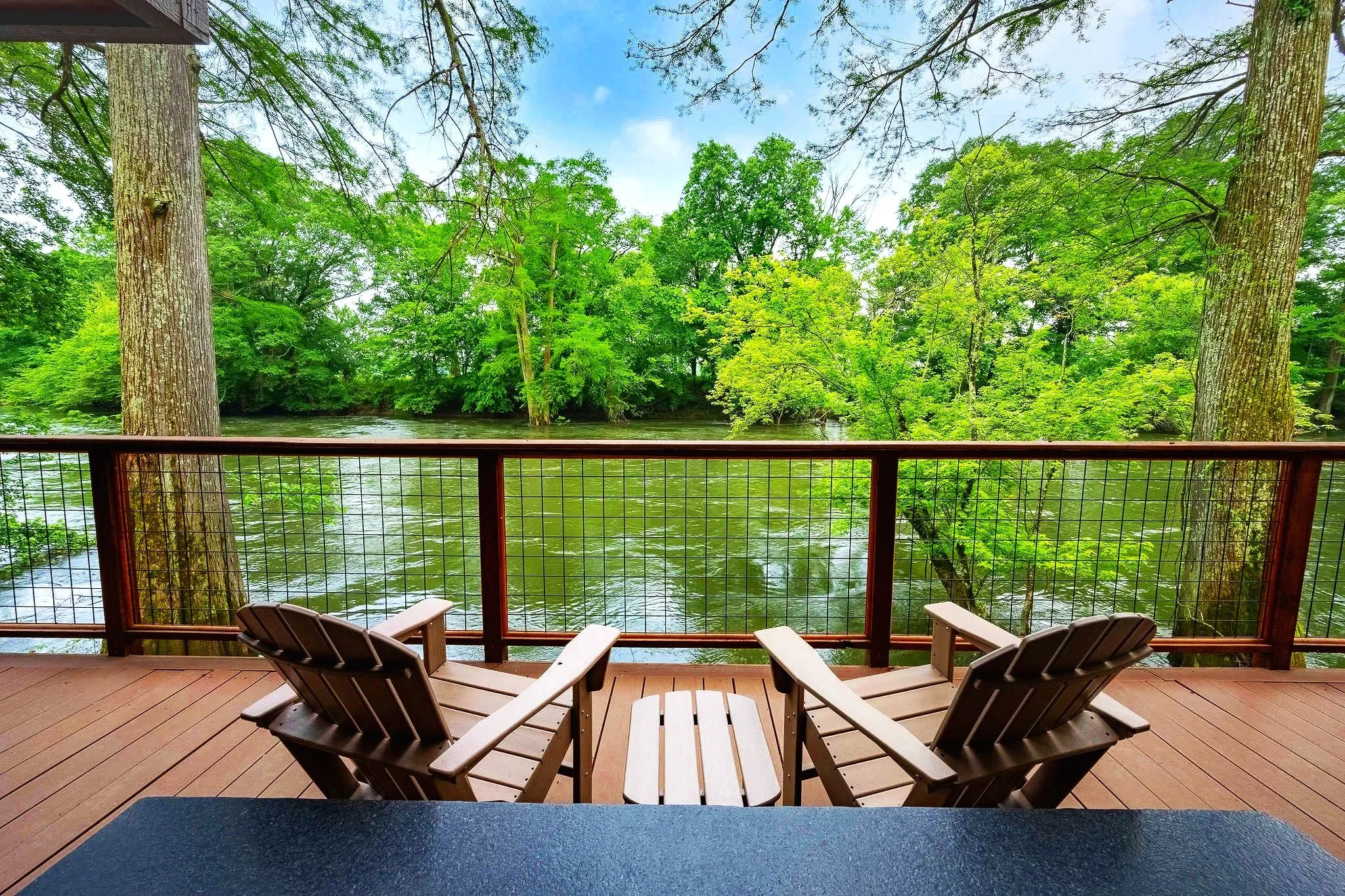 A view from a wooden deck overlooking a river with surrounding lush green trees under a partly cloudy sky.