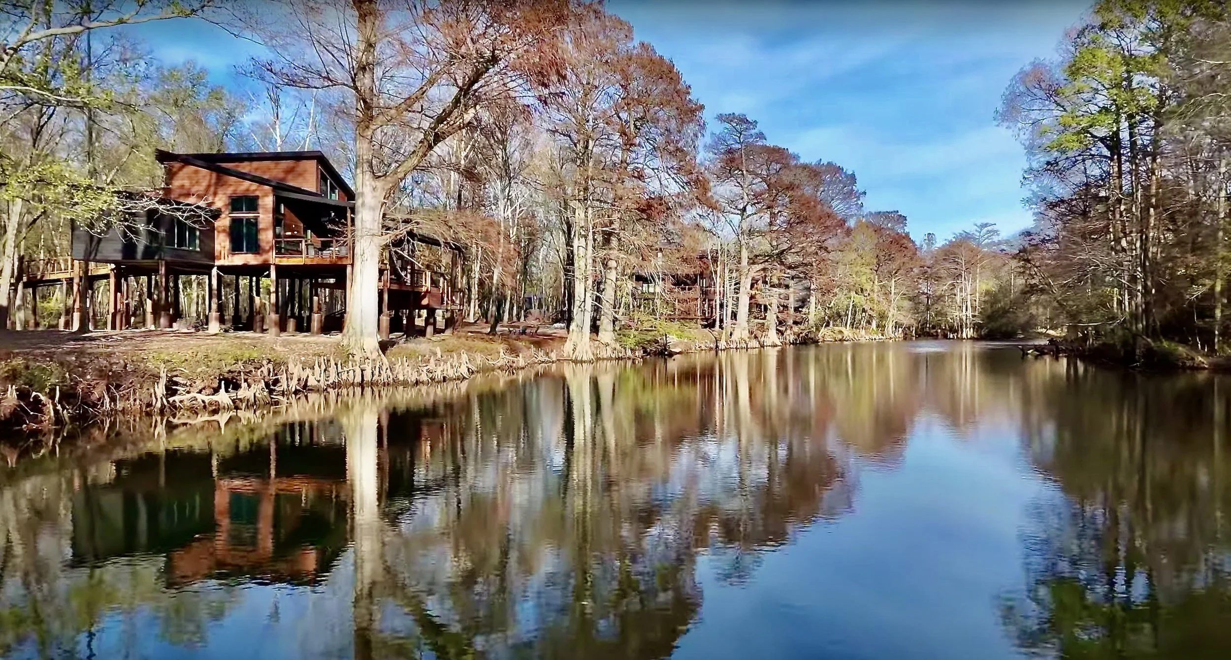 A scenic view of a wooded area with houses built on stilts along a calm river. The trees have autumn leaves, and the sky is blue with some clouds reflected in the water.