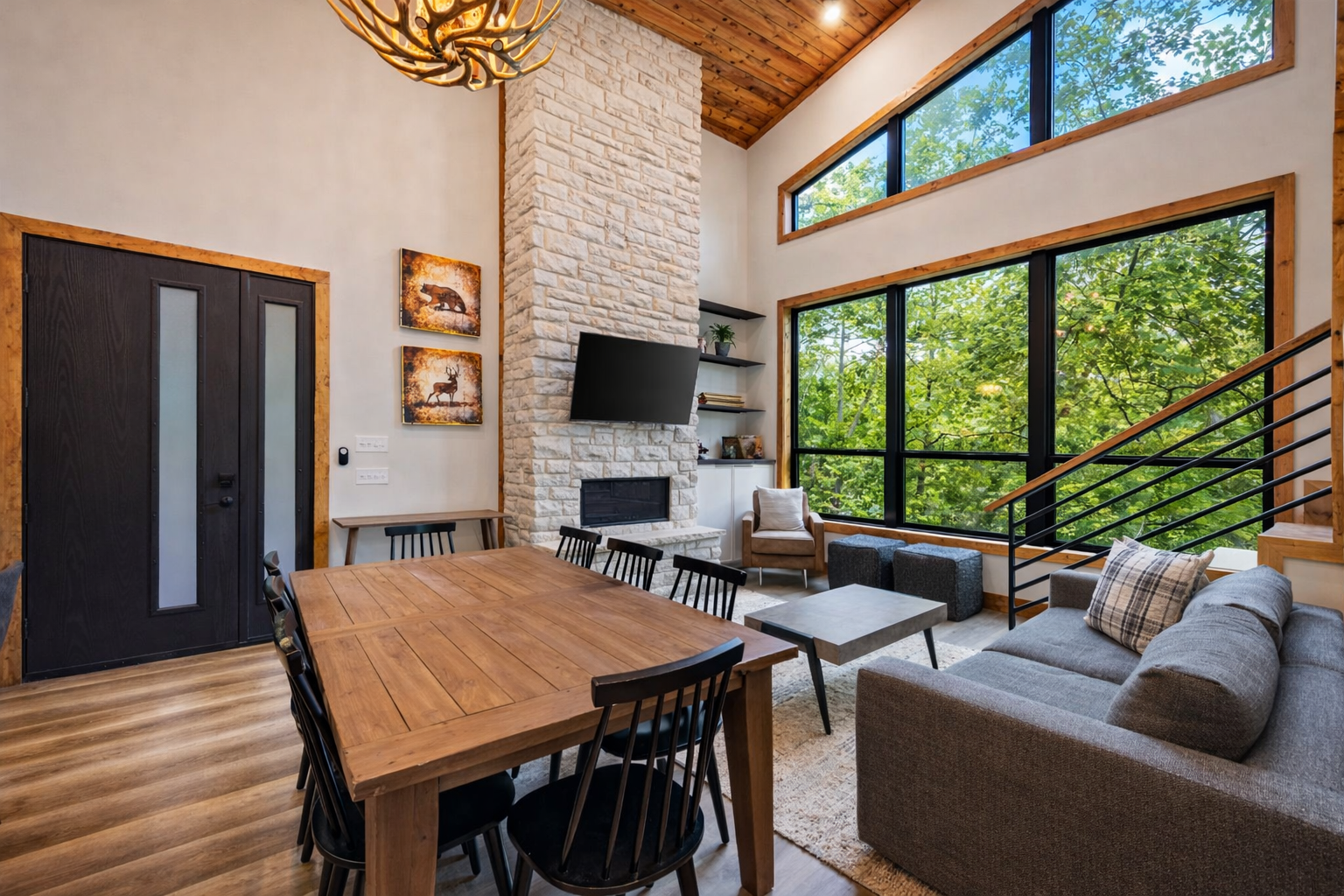 Living room with large front window showing green trees outside, a wooden ceiling, a stone fireplace with a mounted TV, a gray sofa with plaid pillow, coffee tables, and a dining table with black chairs.