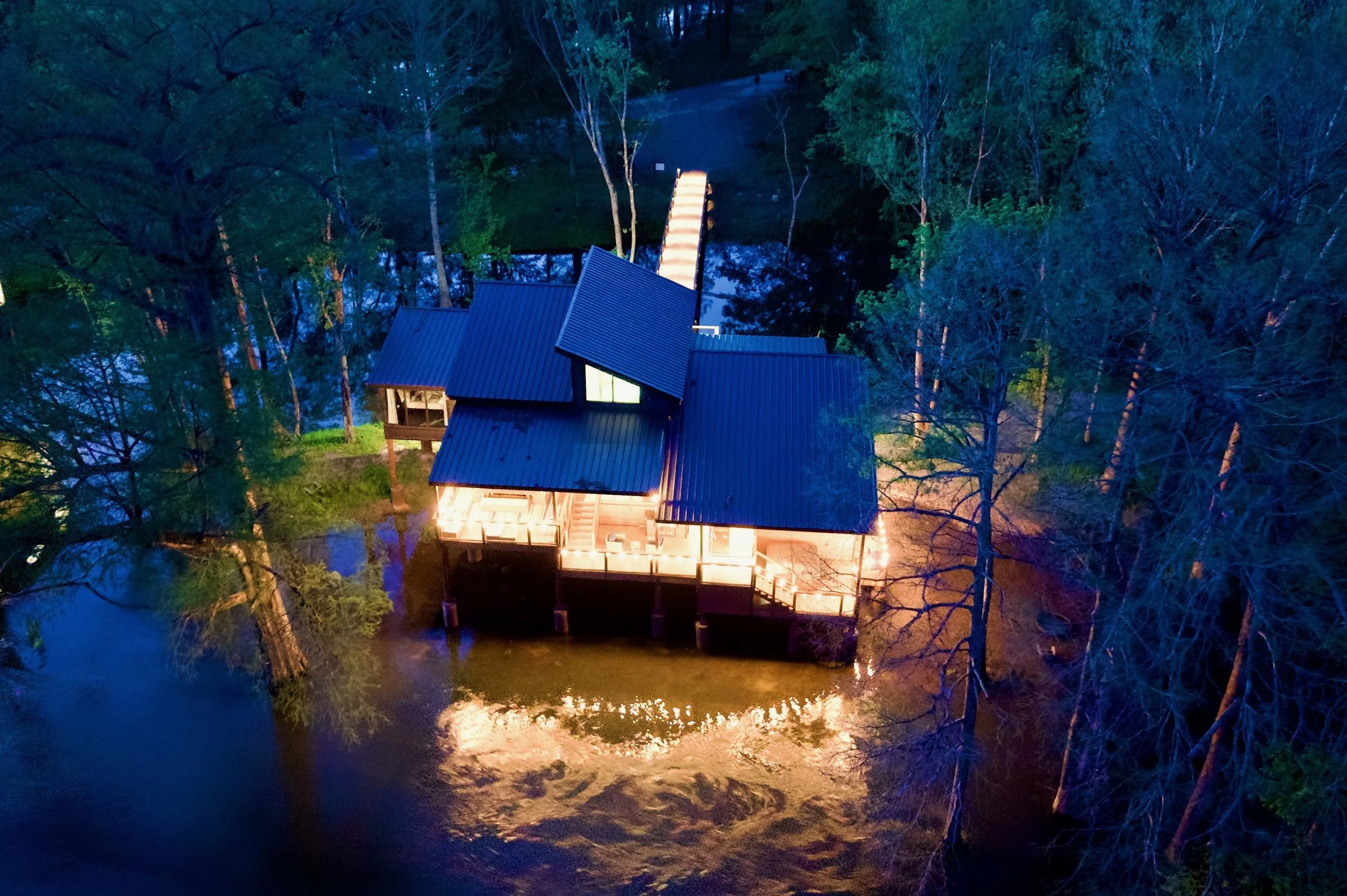 A house with a metal roof on stilts by a river, illuminated at night, surrounded by trees.