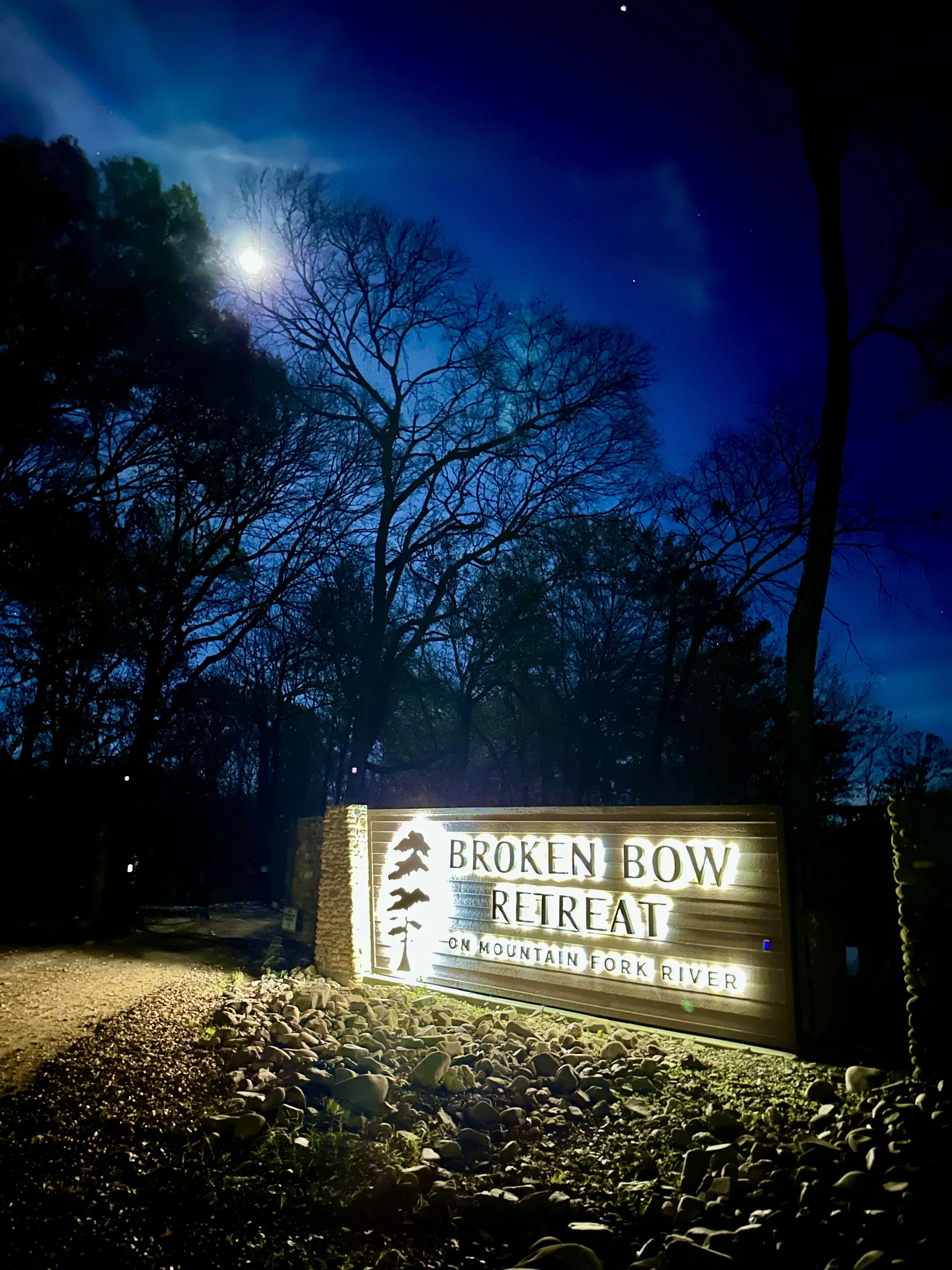 Night view of a sign for Broken Bow Retreat on Mountain Fork River, illuminated with lights, with a wooded area and a bright moon in the sky.