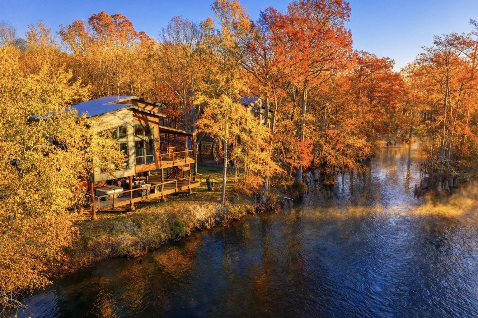 A house by a river during autumn with colorful orange, yellow, and red leaves on the trees, and steam rising from the water in the early morning sunlight.