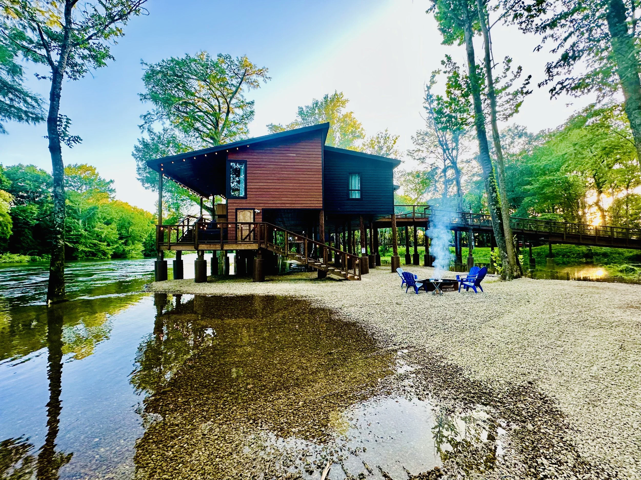A house built on stilts over a river in a forest, with trees and sunlight in the background, and a small fire pit with chairs on the shore.