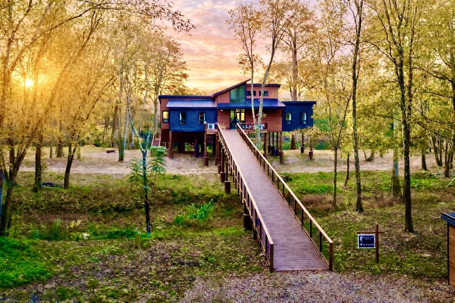 A treehouse with a blue and wooden exterior connected by a long wooden ramp in a forest during sunset with a pink and orange sky.