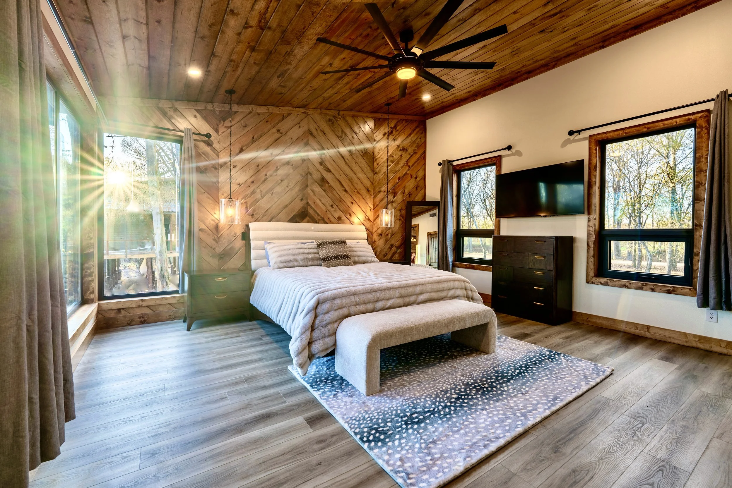 A bedroom with wooden walls and ceiling, featuring a white bed with pillows, a flat-screen TV on a dresser, and large windows with curtains, overlooking trees outside, illuminated by sunlight.